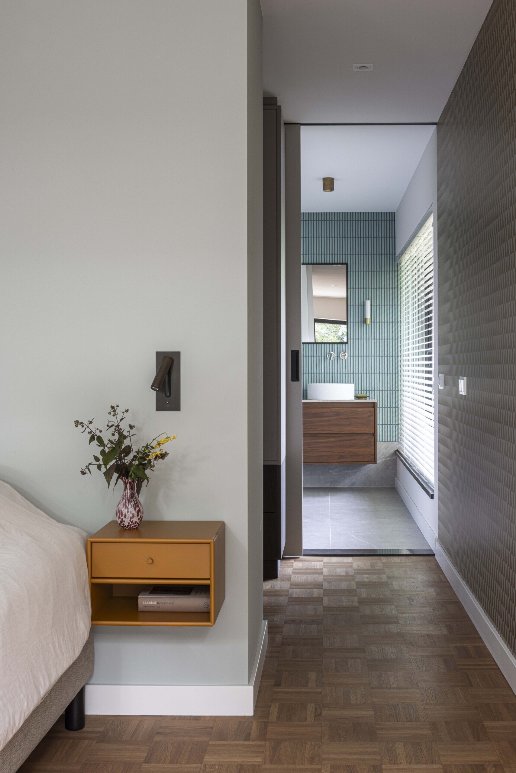 This bedroom showcases the seamless connection between a modern bedroom and a refined en suite bathroom. The space is tied together by warm wood tones and clean, geometric lines, with the parquet flooring guiding the eye from the cozy bedside area into the bathroom.  A wall-mounted mustard yellow nightstand introduces a bold splash of color. Above it, a simple sconce and a vase of freshly picked flowers add a soft, personal touch. The overall effect is calm and considered, with each element carefully chosen to create a sense of quiet sophistication and everyday comfort.