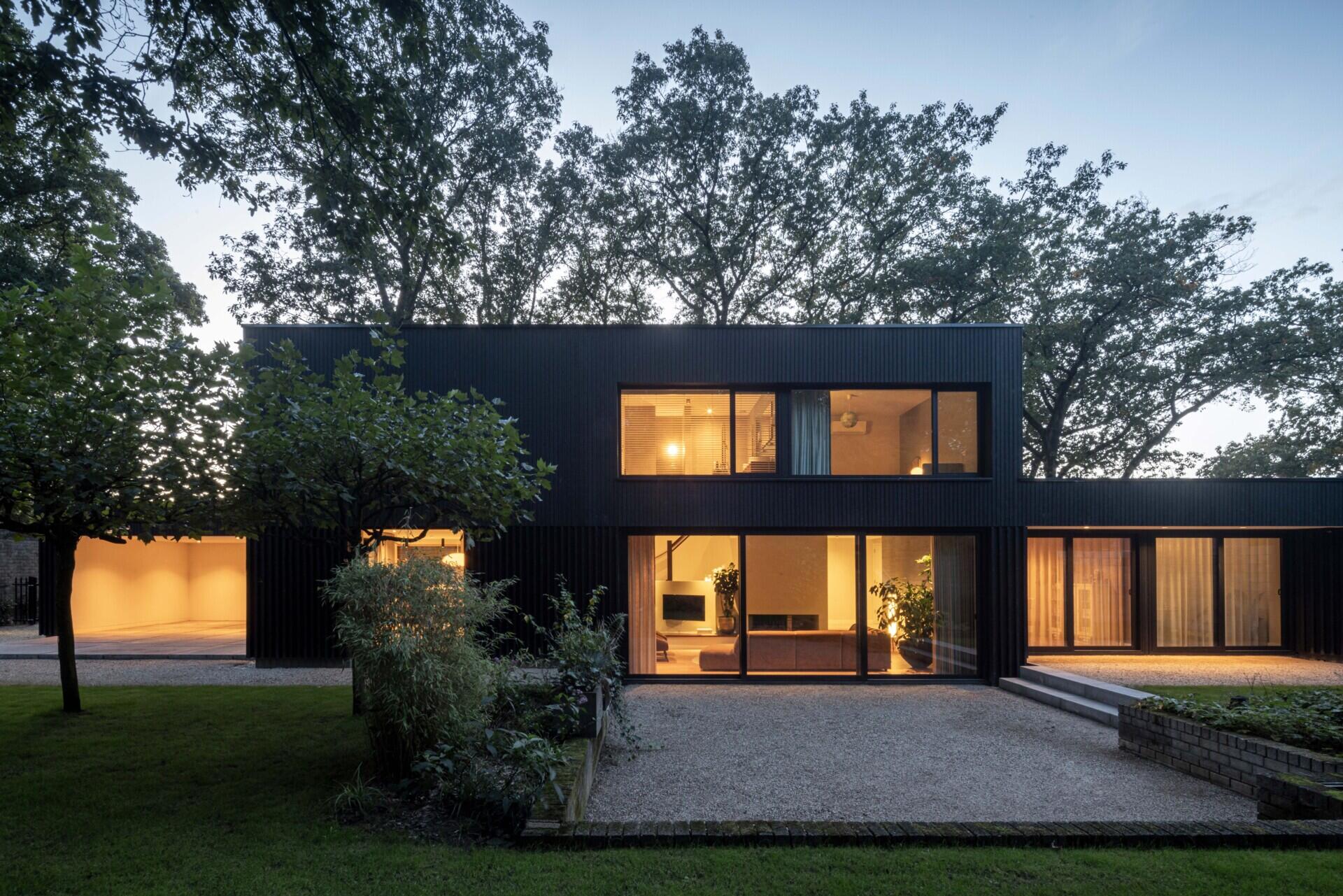 As evening sets in, this modern home reveals its warm, inviting character. Behind the bold silhouette of dark wood cladding, soft interior lighting glows through expansive glass doors and windows, creating a cozy contrast with the surrounding trees.  The layout is open and fluid, with living spaces stretching across the ground floor and private rooms above. Light spills gently onto the gravel paths outside, highlighting the clean architectural lines and the connection between indoors and out. Curtains add privacy without sacrificing the airy, transparent feel.  Set against a backdrop of tall trees and lush greenery, the home feels private yet open, minimal yet welcoming. It’s a perfect example of how thoughtful architecture can feel both modern and timeless when paired with nature.
