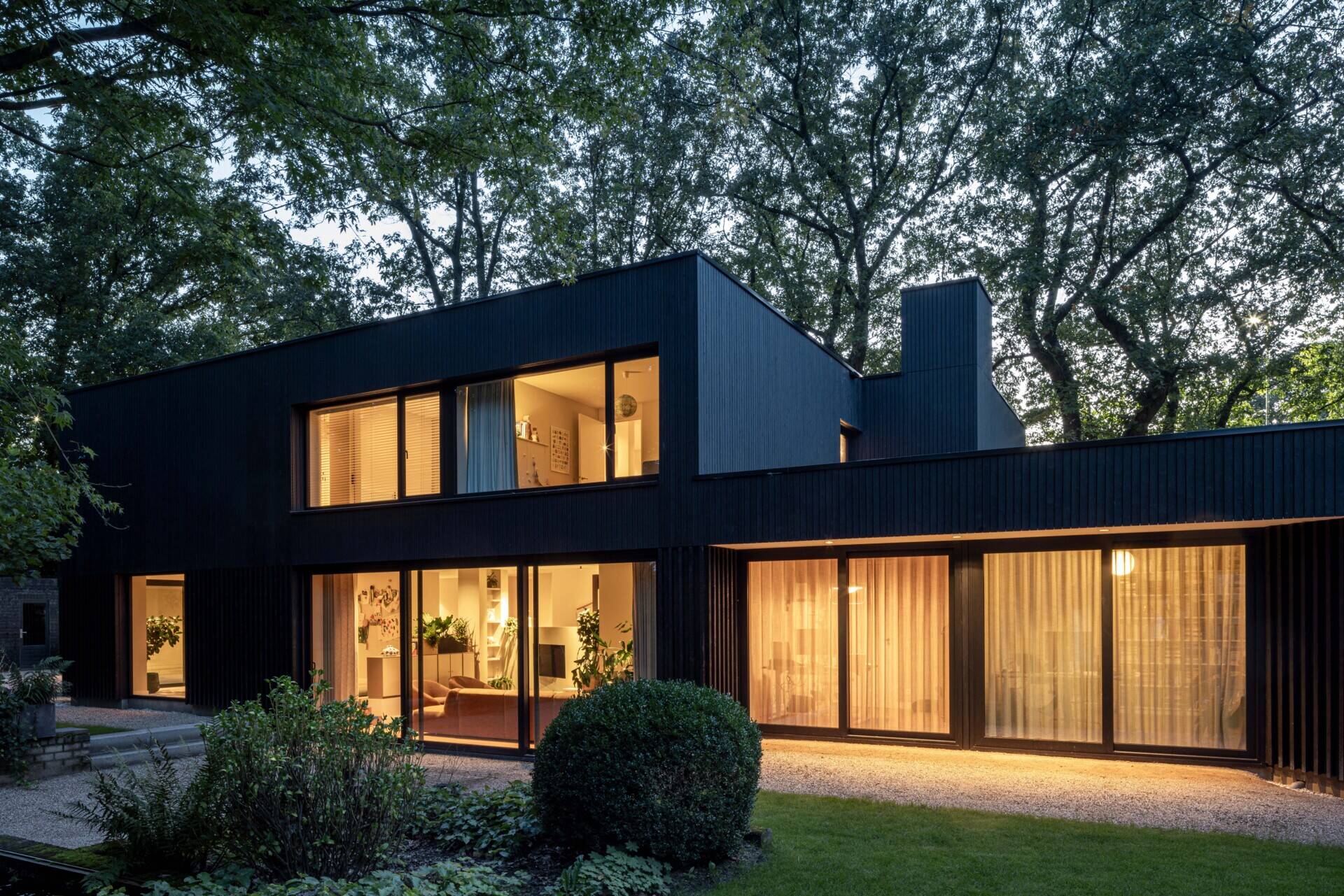 As evening sets in, this modern home reveals its warm, inviting character. Behind the bold silhouette of dark wood cladding, soft interior lighting glows through expansive glass doors and windows, creating a cozy contrast with the surrounding trees.  The layout is open and fluid, with living spaces stretching across the ground floor and private rooms above. Light spills gently onto the gravel paths outside, highlighting the clean architectural lines and the connection between indoors and out. Curtains add privacy without sacrificing the airy, transparent feel.  Set against a backdrop of tall trees and lush greenery, the home feels private yet open, minimal yet welcoming. It’s a perfect example of how thoughtful architecture can feel both modern and timeless when paired with nature.