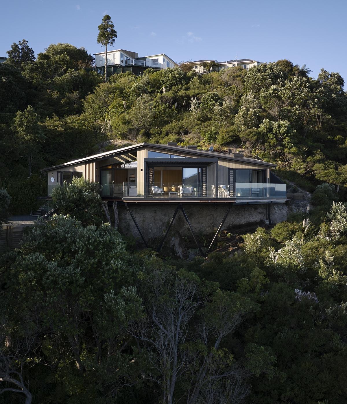 Perched high above the coastline, this striking modern home blends seamlessly into the lush hillside. Cantilevered over the cliff on slender steel supports, it features wraparound glass balustrade decks, warm timber cladding, and floor-to-ceiling glazing that frames panoramic views of the sea and sky. A bold, nature-integrated design that feels like a luxury treehouse.