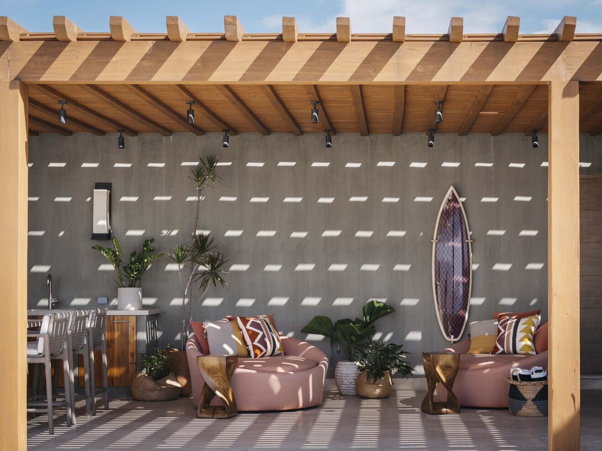 This outdoor lounge captures that perfect blend of sun-drenched and shaded, where the light filters through the slatted roof in a mesmerizing dance across every surface. The wood pergola feels warm and welcoming, casting patterned shadows that change with the day. A pair of circular loungers in soft desert rose invite you to sink in and stay awhile, their colorful pillows echoing the tones of clay, sand, and sunset. A surfboard-turned-art piece adds a playful, coastal energy to the concrete wall, while lush potted plants soften the space with their broad green leaves.