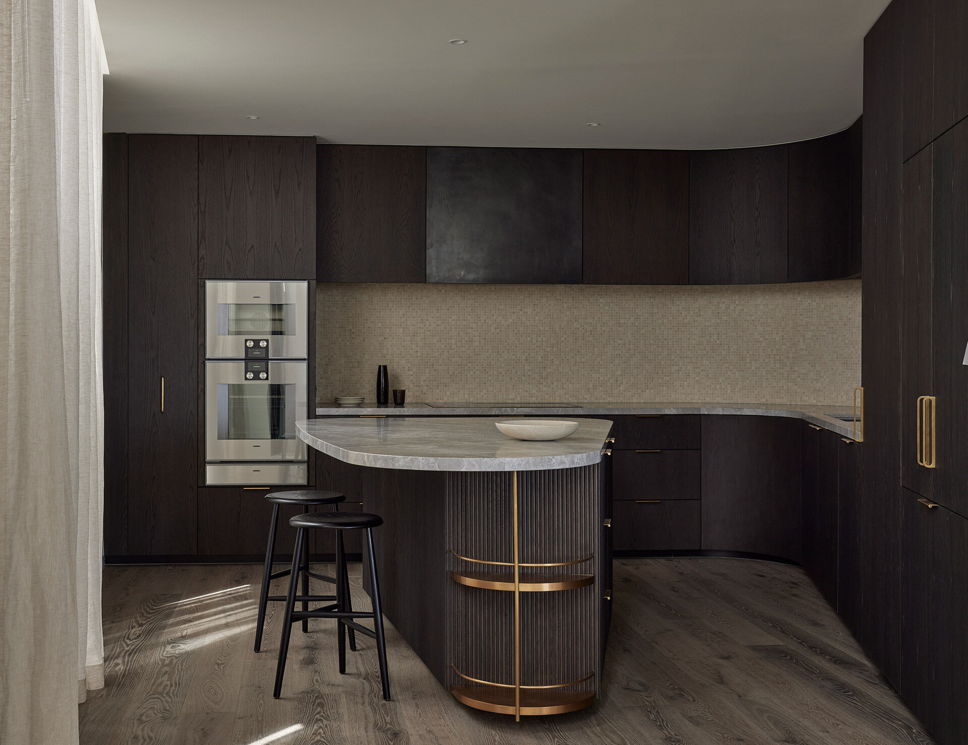 In this kitchen, dark timber joinery wraps the space in warmth, while softly curved cabinetry and brushed gold hardware add a sense of sculptural elegance. A fluted kitchen island with stone benchtop anchors the room, paired with sleek black bar stools that keep the palette grounded.