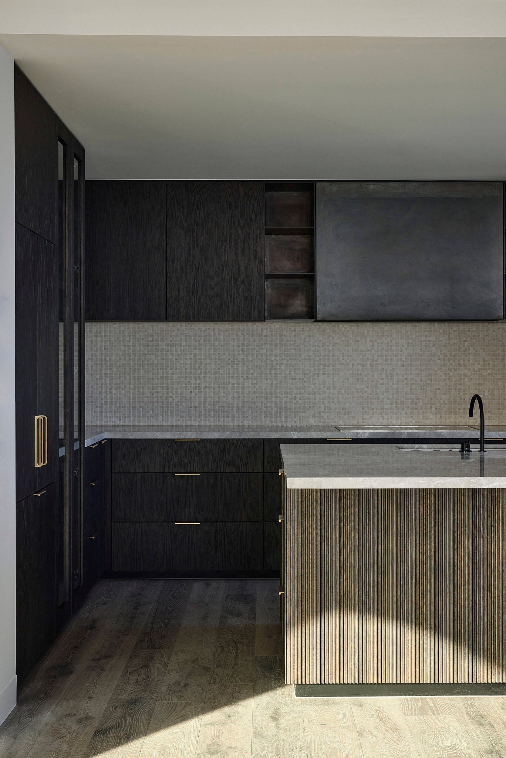 In this kitchen, dark timber joinery wraps the space in warmth, while softly curved cabinetry and brushed gold hardware add a sense of sculptural elegance. A fluted kitchen island with stone benchtop anchors the room, paired with sleek black bar stools that keep the palette grounded.
