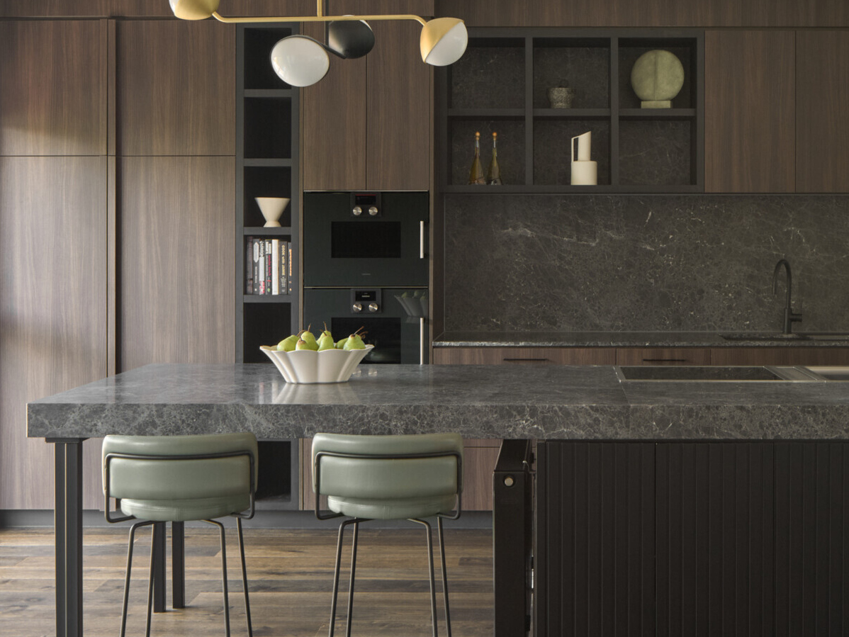 In this kitchen, materials were chosen for both their beauty and their durability. Dark timber cabinetry and natural stone surfaces bring warmth and texture, while sculptural pendant lights introduce contrast and a sense of artfulness. The island functions as both a prep space and a place to gather. Seating is cleverly integrated at both ends: one side extends into a casual dining area with a black table and matching chairs, while the other side features bar stools for a more relaxed perch. This dual arrangement brings flexibility to everyday life and entertaining.