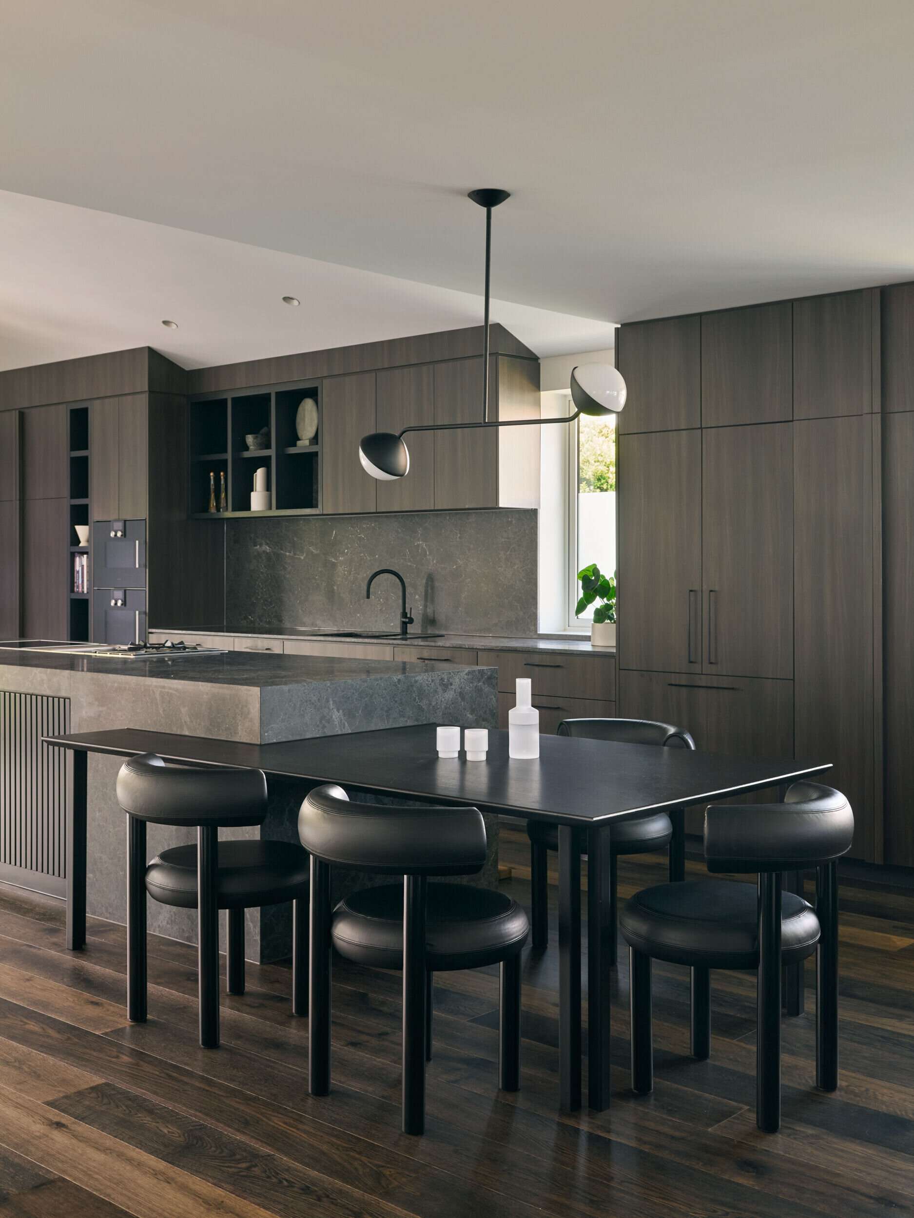 In this kitchen, materials were chosen for both their beauty and their durability. Dark timber cabinetry and natural stone surfaces bring warmth and texture, while sculptural pendant lights introduce contrast and a sense of artfulness. The island functions as both a prep space and a place to gather. Seating is cleverly integrated at both ends: one side extends into a casual dining area with a black table and matching chairs, while the other side features bar stools for a more relaxed perch. This dual arrangement brings flexibility to everyday life and entertaining.