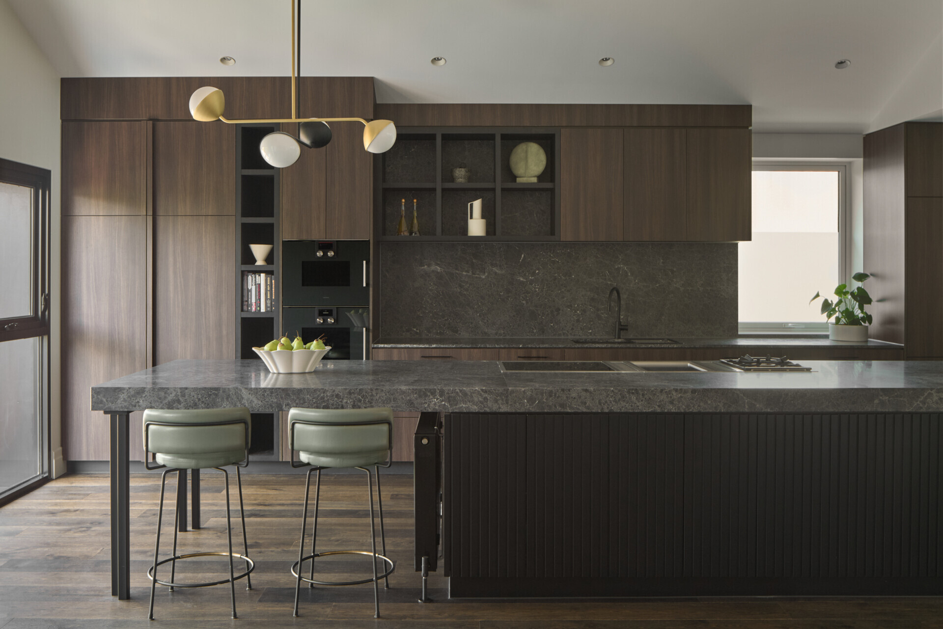 In this kitchen, materials were chosen for both their beauty and their durability. Dark timber cabinetry and natural stone surfaces bring warmth and texture, while sculptural pendant lights introduce contrast and a sense of artfulness. The island functions as both a prep space and a place to gather. Seating is cleverly integrated at both ends: one side extends into a casual dining area with a black table and matching chairs, while the other side features bar stools for a more relaxed perch. This dual arrangement brings flexibility to everyday life and entertaining.