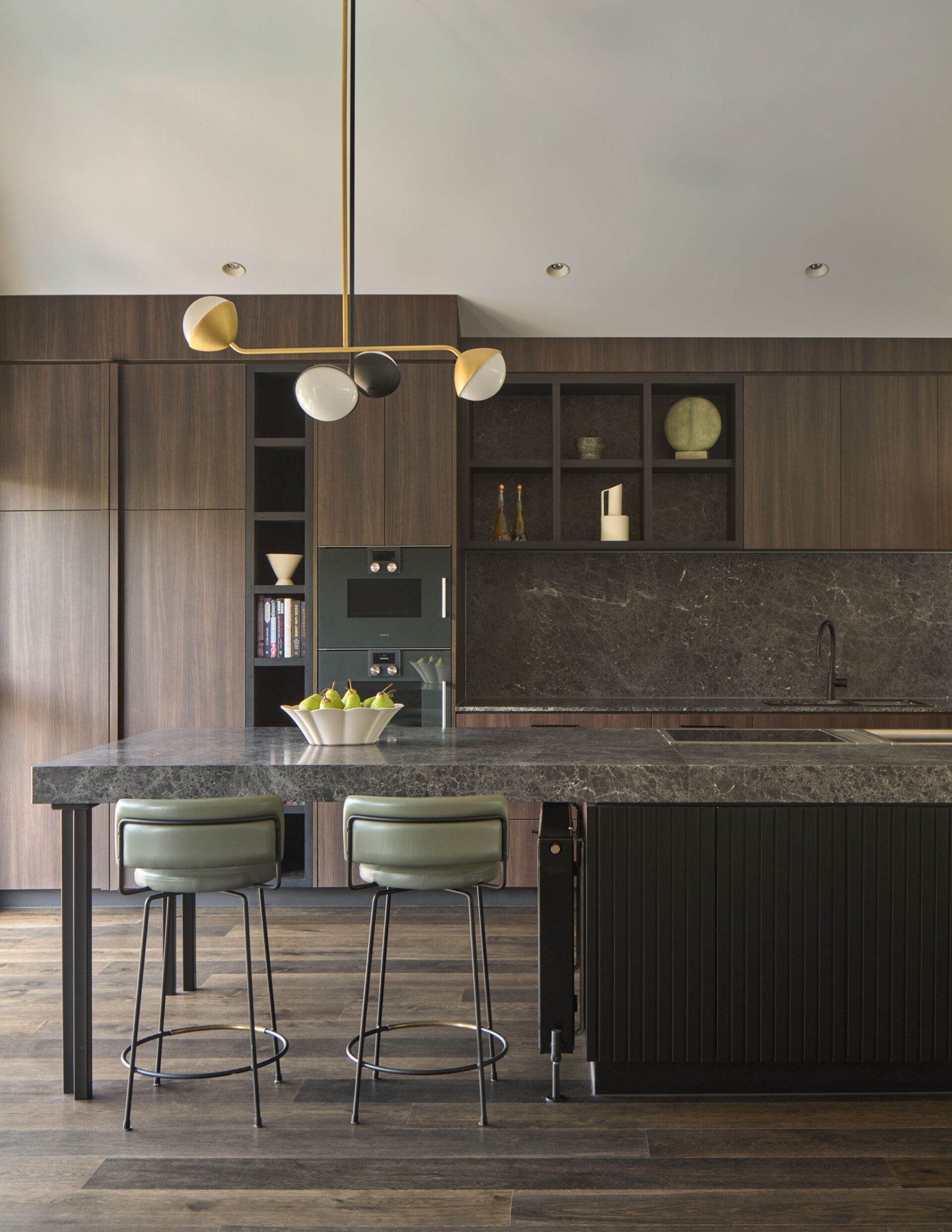In this kitchen, materials were chosen for both their beauty and their durability. Dark timber cabinetry and natural stone surfaces bring warmth and texture, while sculptural pendant lights introduce contrast and a sense of artfulness. The island functions as both a prep space and a place to gather. Seating is cleverly integrated at both ends: one side extends into a casual dining area with a black table and matching chairs, while the other side features bar stools for a more relaxed perch. This dual arrangement brings flexibility to everyday life and entertaining.