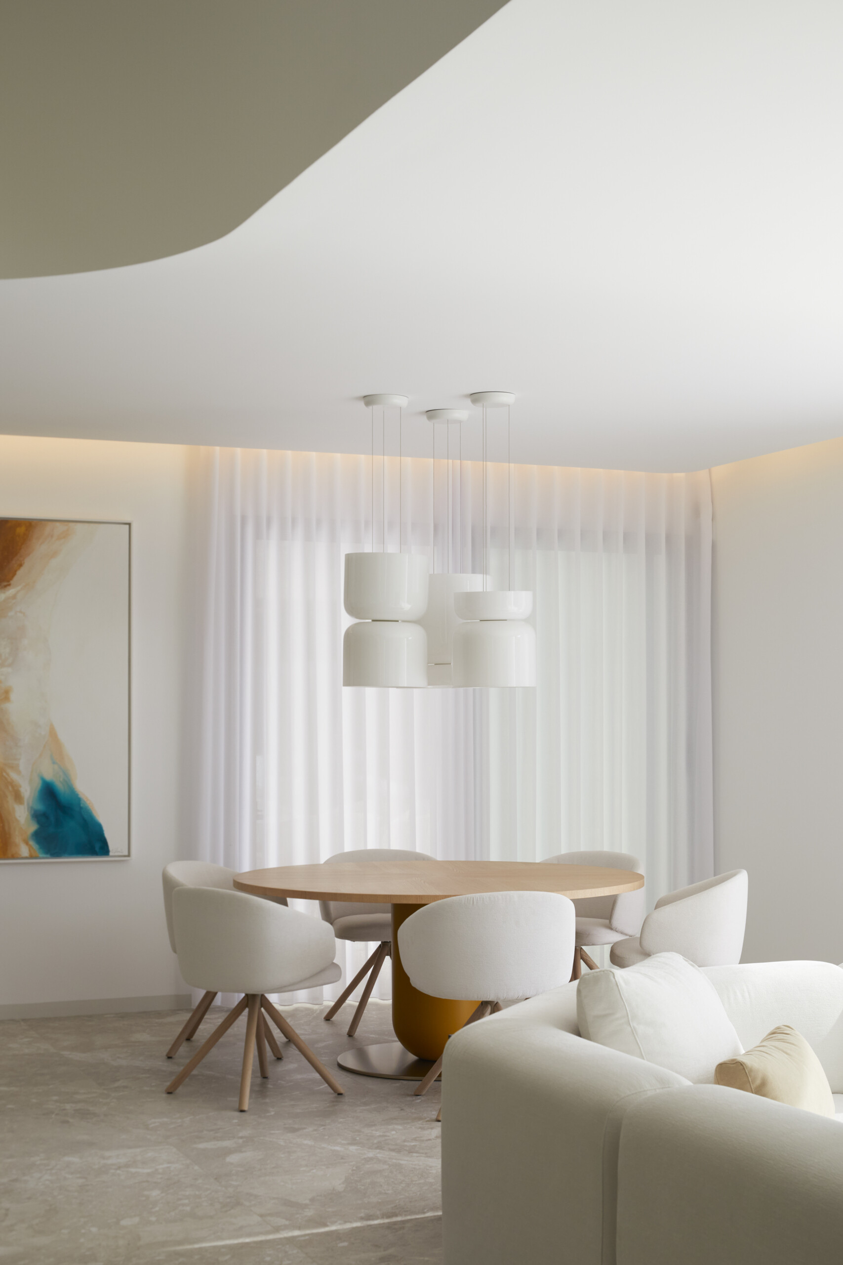 A round wood table sits at the heart of the space, anchored by a golden ochre base that adds just a hint of playfulness beneath its smooth surface. Surrounding it, plush cream chairs with curved backs echo the gentle shapes found throughout the home, offering both comfort and style. Above, a sculptural cluster of white pendant lights floats like a modern mobile, drawing the eye upward without overwhelming the room. 