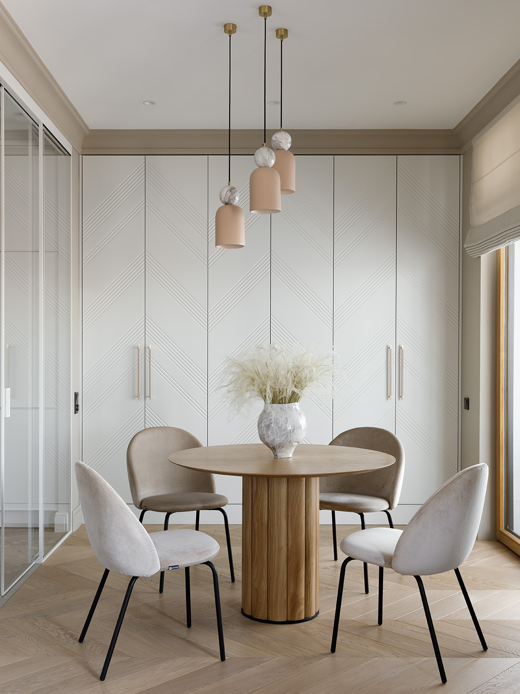 Soft neutrals and layered textures create a serene dining nook. A round wood table adds warmth, while curved chairs invite lingering. Pale pendants with marble accents bring a touch of whimsy, and subtly patterned cabinet doors add texture to the space.