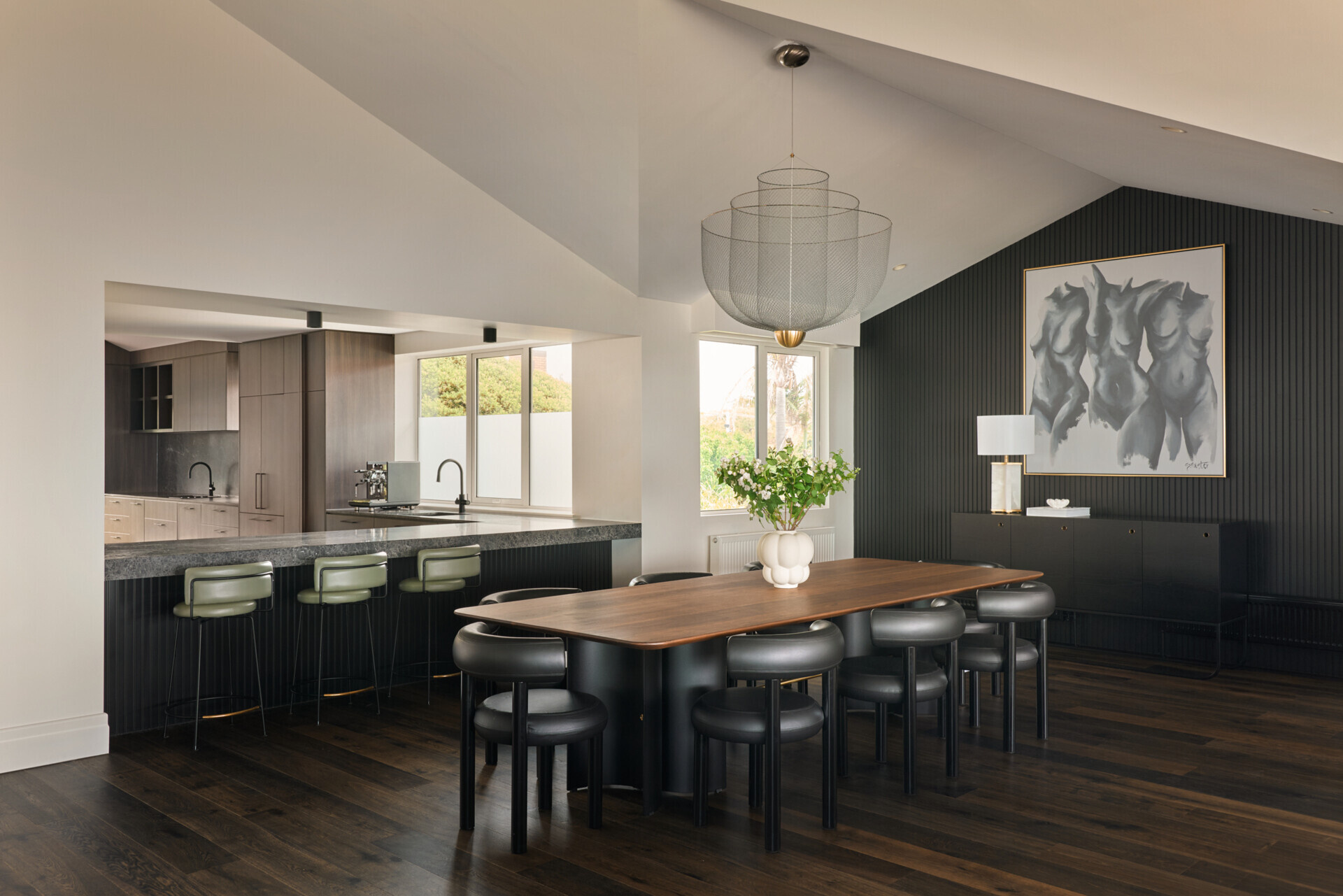 The dining room, with Art Deco references, balances drama and warmth with a rich, tonal palette. A long timber table and curved black chairs anchor the space, while a sculptural pendant adds softness overhead. Vertical black paneling and a bold artwork create contrast, framed by natural light from large windows. It's a space that feels both elegant and relaxed, perfect for gatherings or quiet meals. Above, a tiered wire-frame pendant light adds visual softness and draws attention to the pitched ceiling without dominating the room.