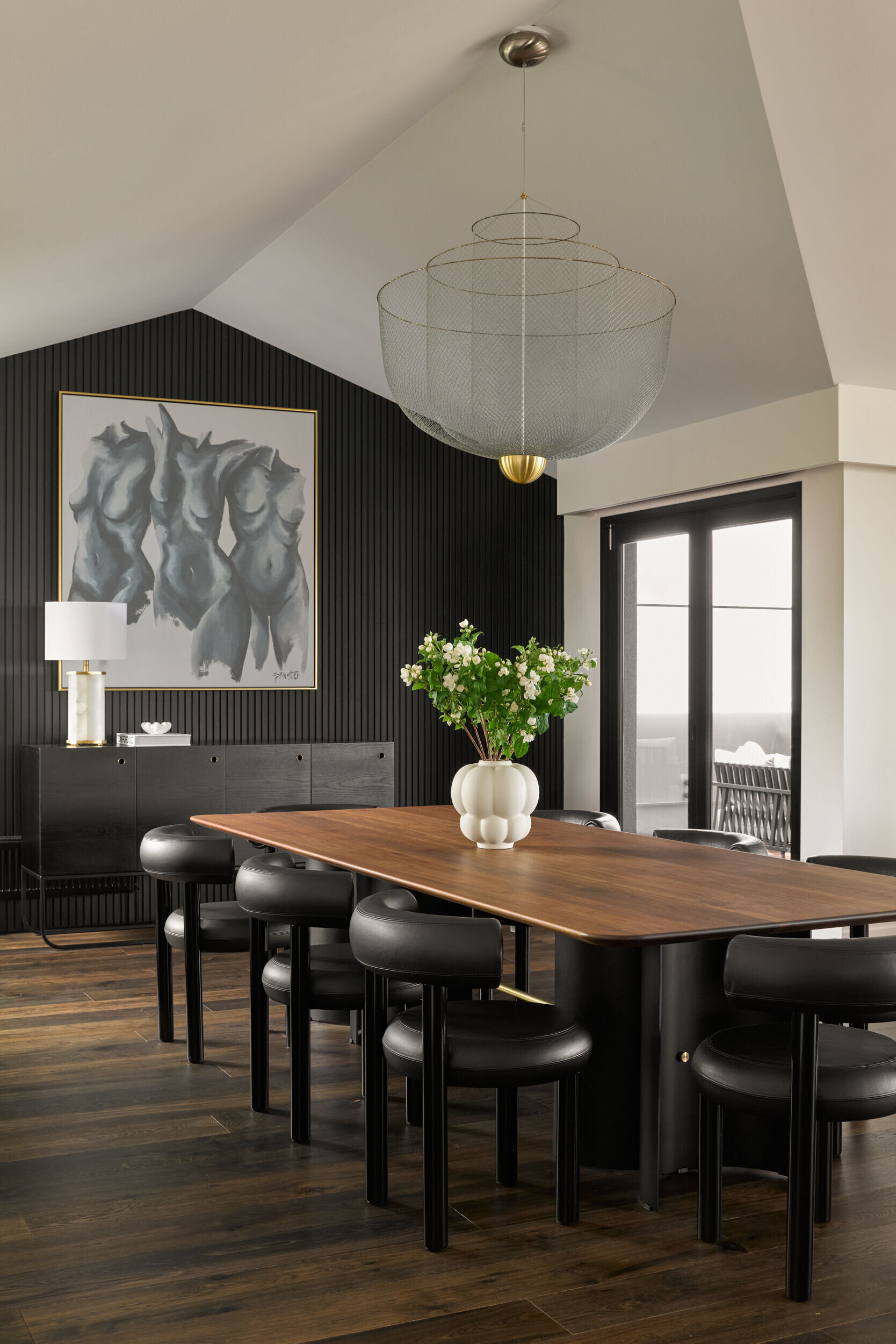 The dining room, with Art Deco references, balances drama and warmth with a rich, tonal palette. A long timber table and curved black chairs anchor the space, while a sculptural pendant adds softness overhead. Vertical black paneling and a bold artwork create contrast, framed by natural light from large windows. It's a space that feels both elegant and relaxed, perfect for gatherings or quiet meals. Above, a tiered wire-frame pendant light adds visual softness and draws attention to the pitched ceiling without dominating the room.