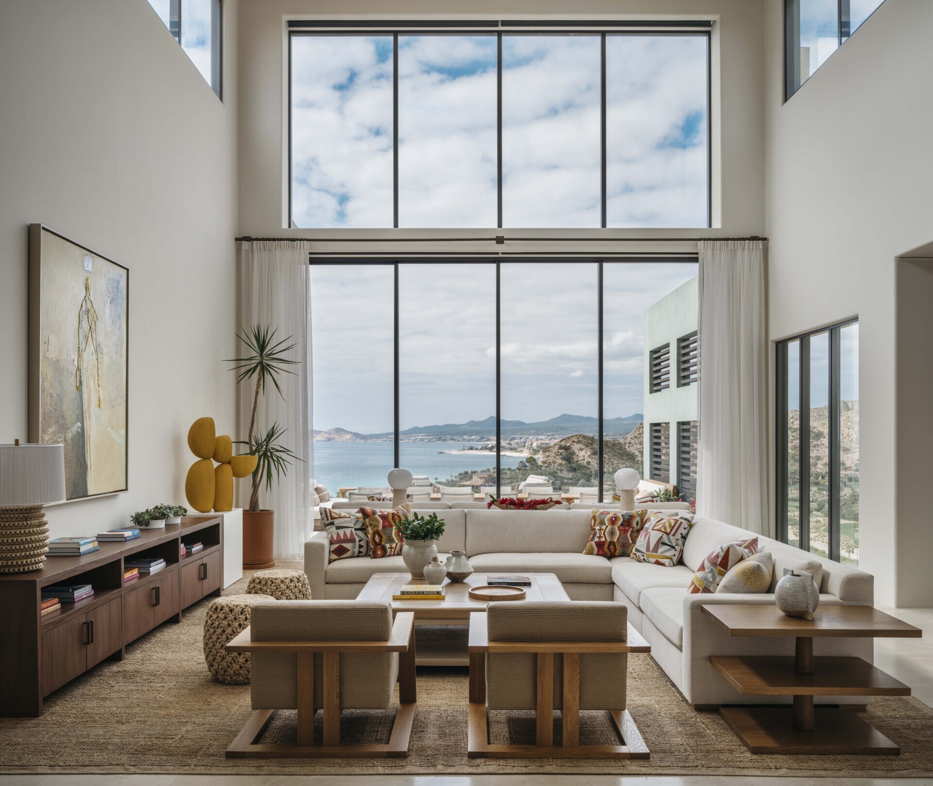 The living room is a study in contrast, its neutral, modern shell becomes a gallery for rich textiles, collected art, and sculptural lighting. Anchored by a bold Frank Arnold painting sourced locally in San Jose del Cabo, the space welcomes conversation and lounging in equal measure. Natural textures like seagrass and rope weave through ottomans and rugs, grounding the room in regional craft traditions. Soft, neutral-toned upholstery allows the vivid accents to take center stage, making the living space feel layered yet light.