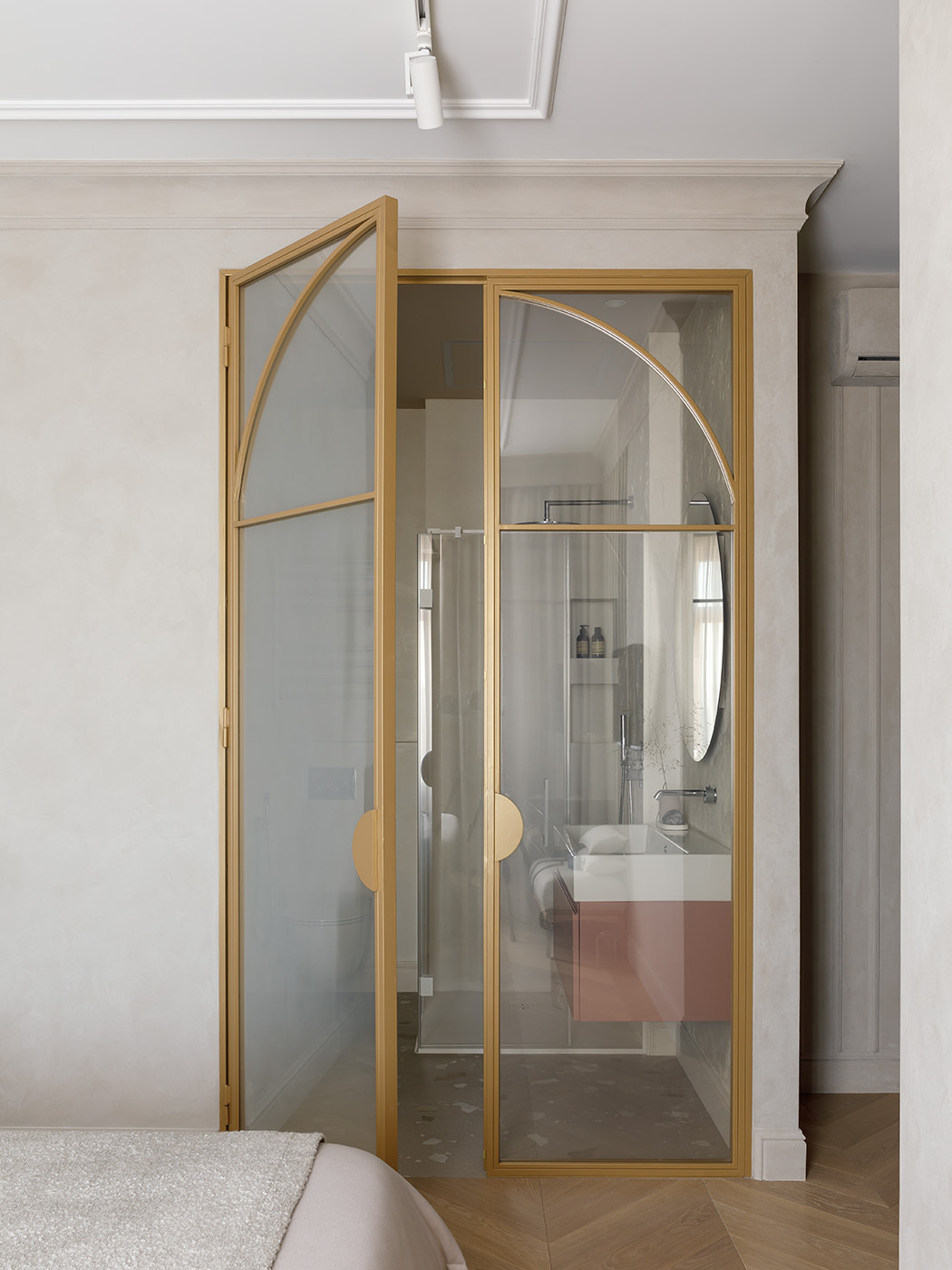 A pair of arched glass doors framed in brushed gold instantly elevate this ensuite bathroom, turning a simple transition into a quiet statement. Soft, sandy walls and pale wood flooring create a seamless flow from bedroom to bath, while the terracotta vanity brings just a touch of grounded color.