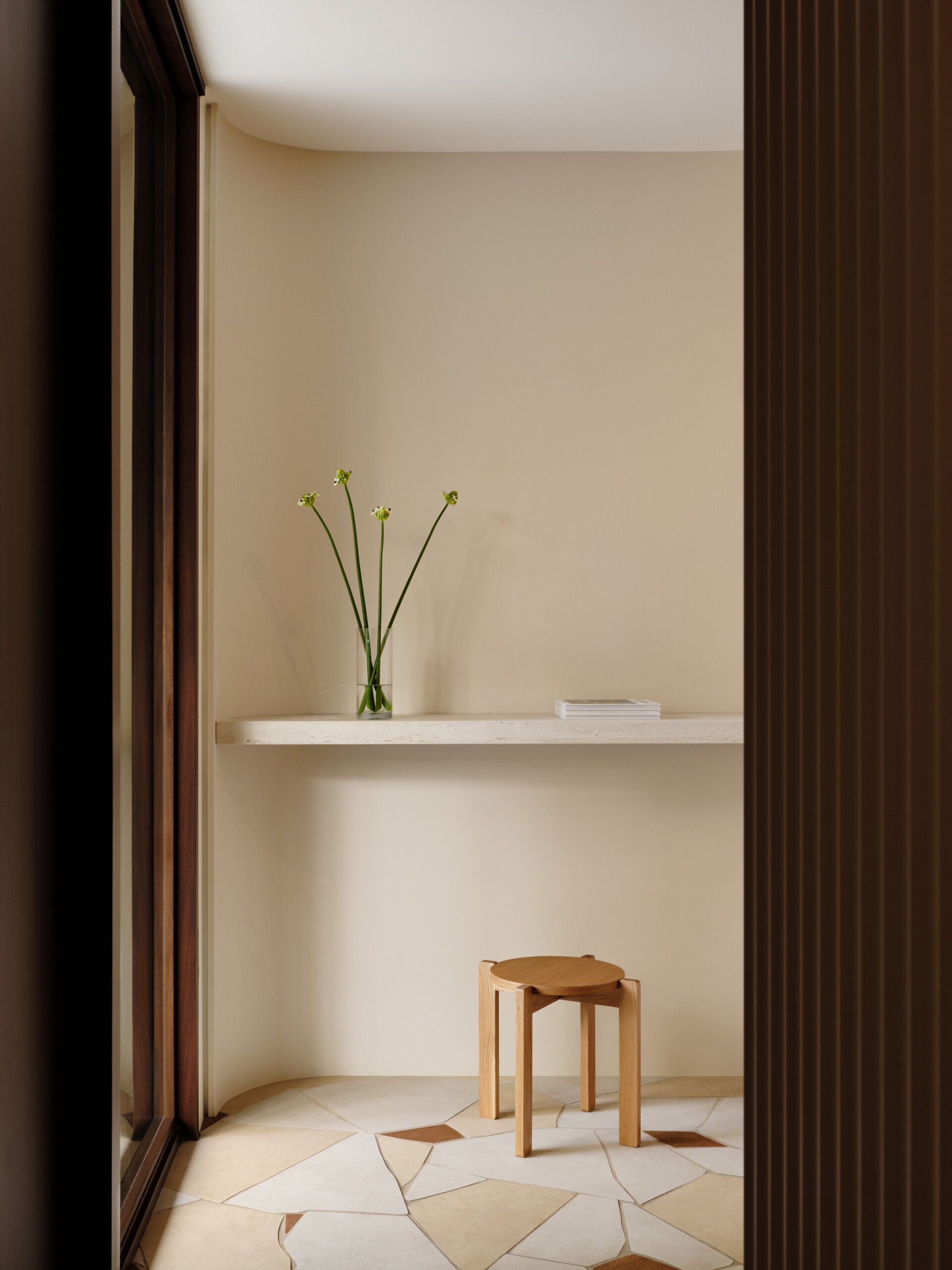 The entryway offers a quiet study in restraint and beauty. A simple floating shelf in pale stone runs along a softly curved wall, echoing the gentle geometry found throughout the home. The styling is minimal yet poetic—a slender glass vase with delicate green-stemmed blooms brings life to the space without disturbing its calm.  Below, the floor steals a quiet moment of attention. Laid in an opus incertum pattern, the ceramic tiles resemble irregular shards of natural slate, with tones of cream, sand, and terracotta that add depth and warmth. A low, sculptural stool in natural oak sits alone, its softly rounded edges mirroring the architectural gestures found throughout the house.  This small space captures the essence of the entire home: quiet elegance, natural light, and a deep respect for materiality.