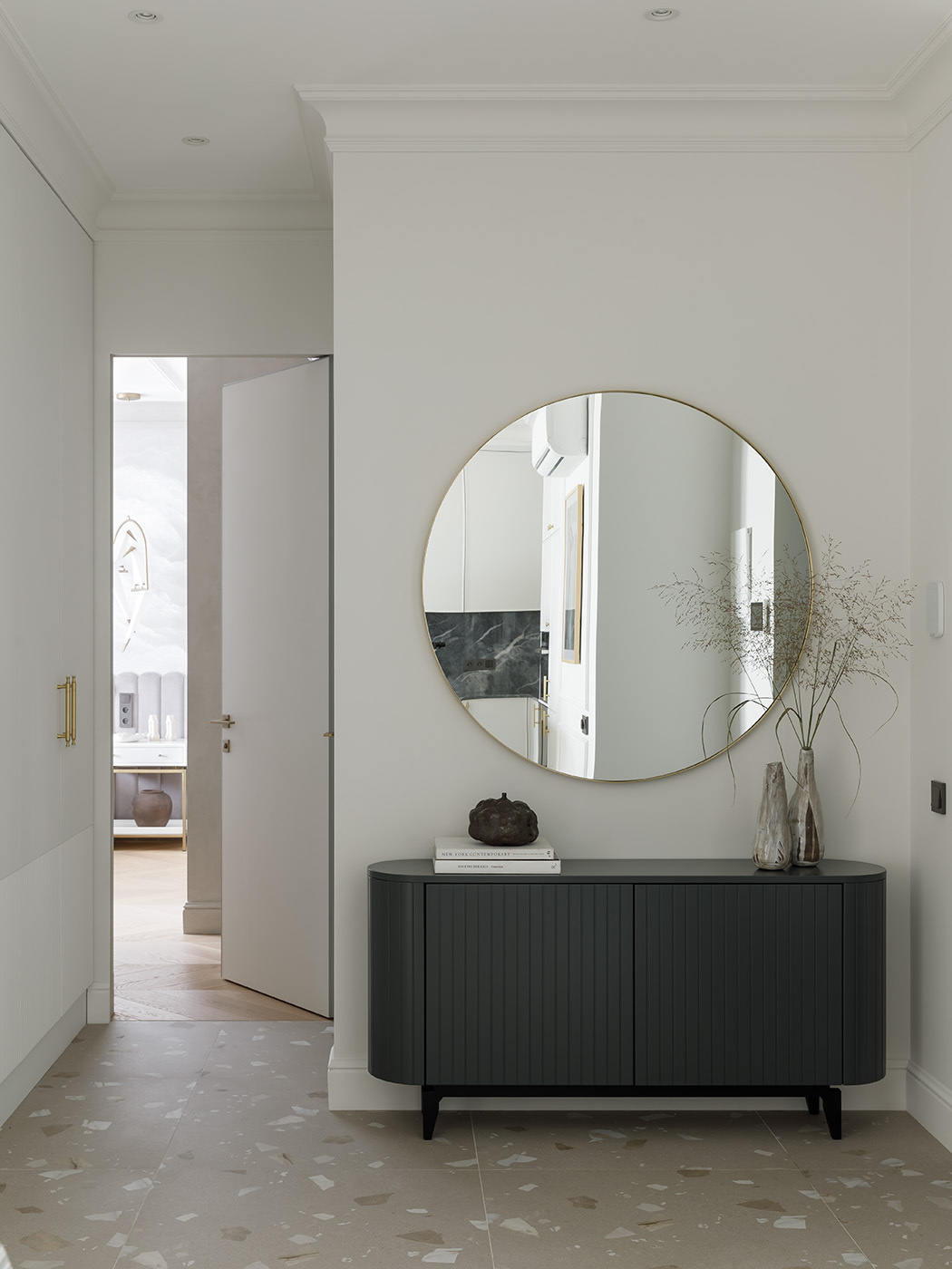 This entryway feels calm and confident, with just the right mix of texture and polish. The dark console adds depth and contrast against soft white walls, while its fluted design gives a quiet rhythm to the space. A round mirror in a thin gold frame reflects light and subtly opens up the space.