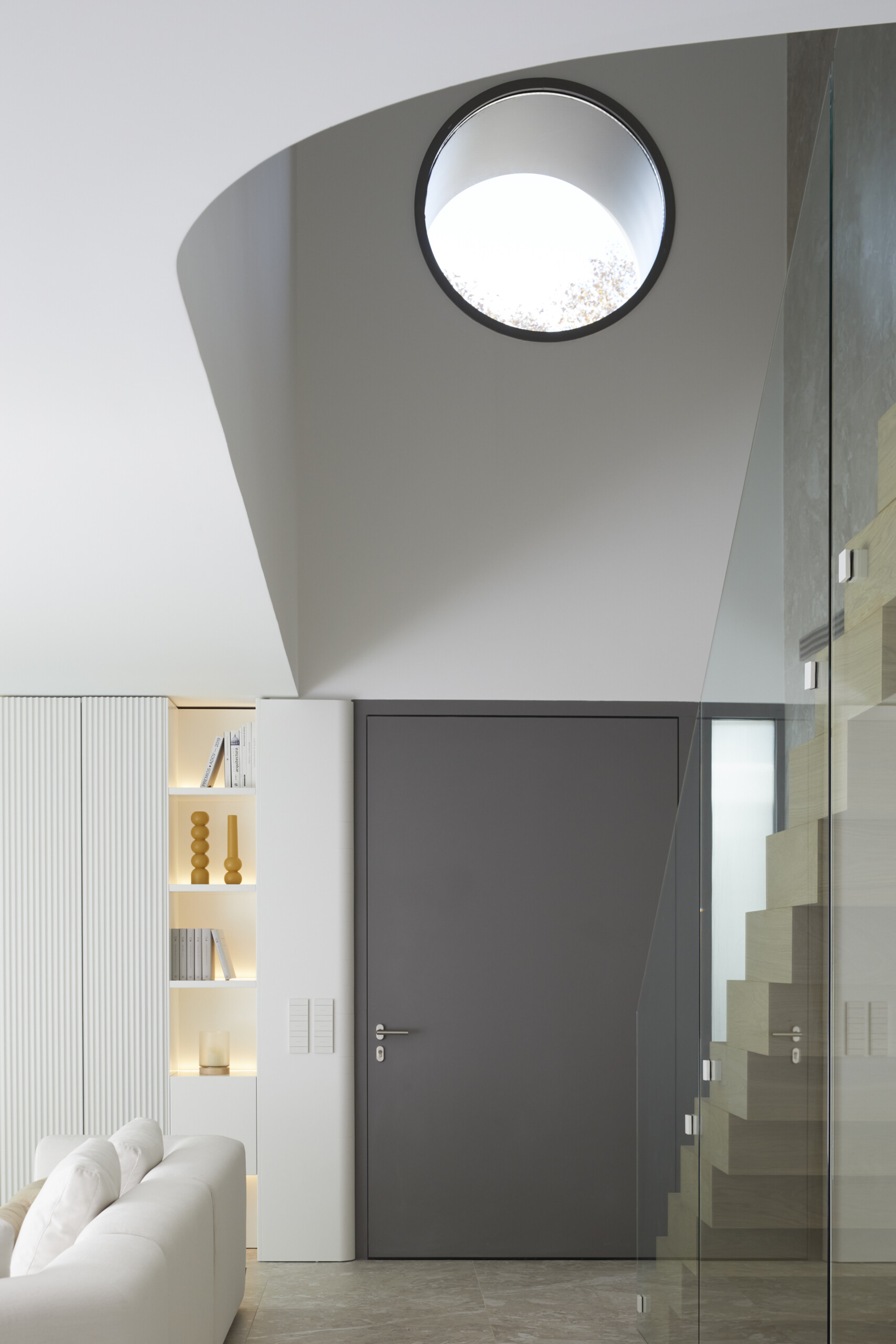  The entryway pairs architectural drama with quiet restraint. A tall charcoal door sits beneath a sculptural ceiling and round skylight, while built-in shelves and natural finishes offer a warm, curated welcome.