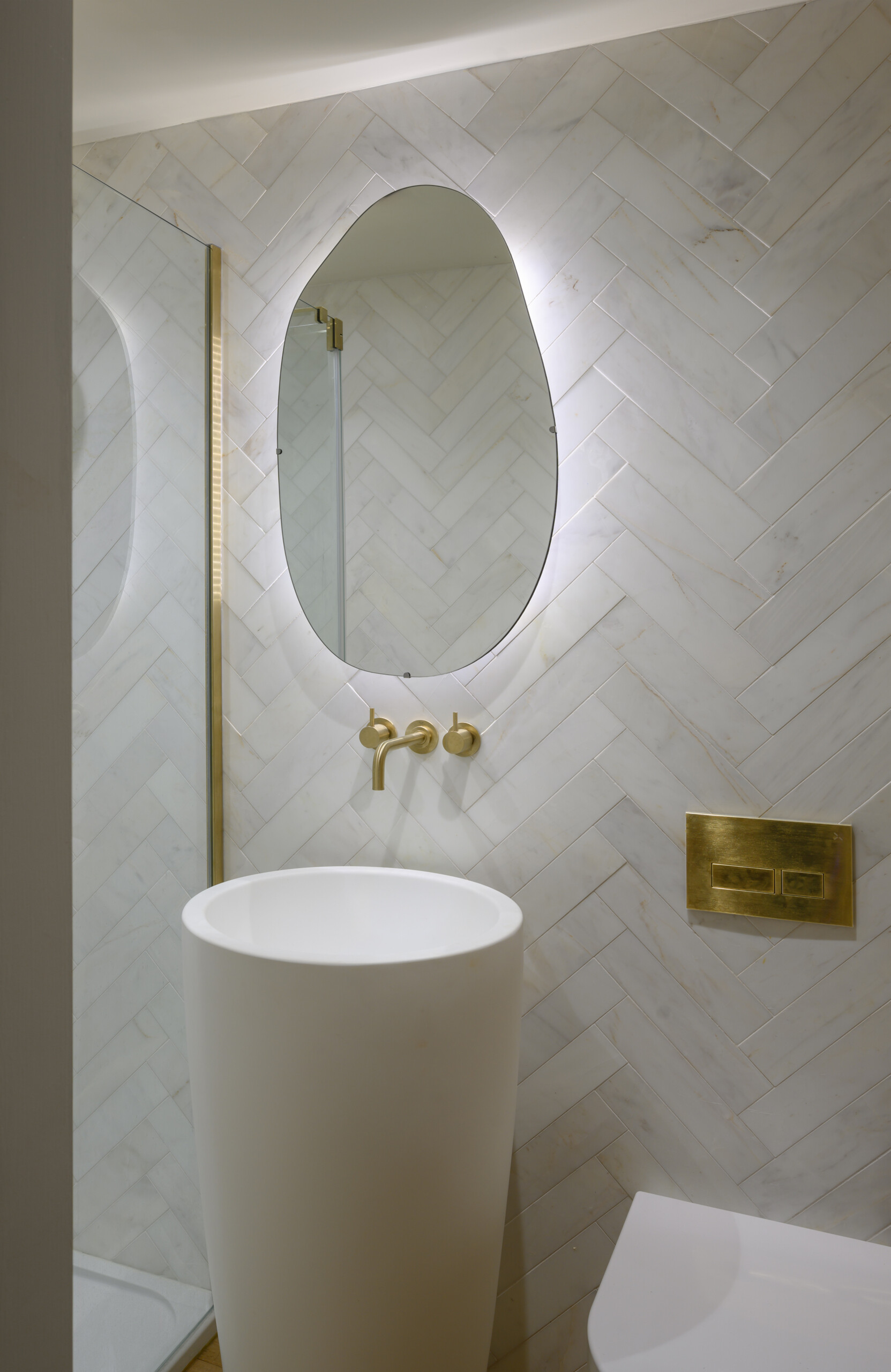 In this guest bathroom, pale stone tiles are laid in a herringbone pattern across the walls, creating soft texture and visual interest. A freestanding cylindrical basin sits beneath an organically shaped mirror, while brass details give the space a refined, contemporary edge.