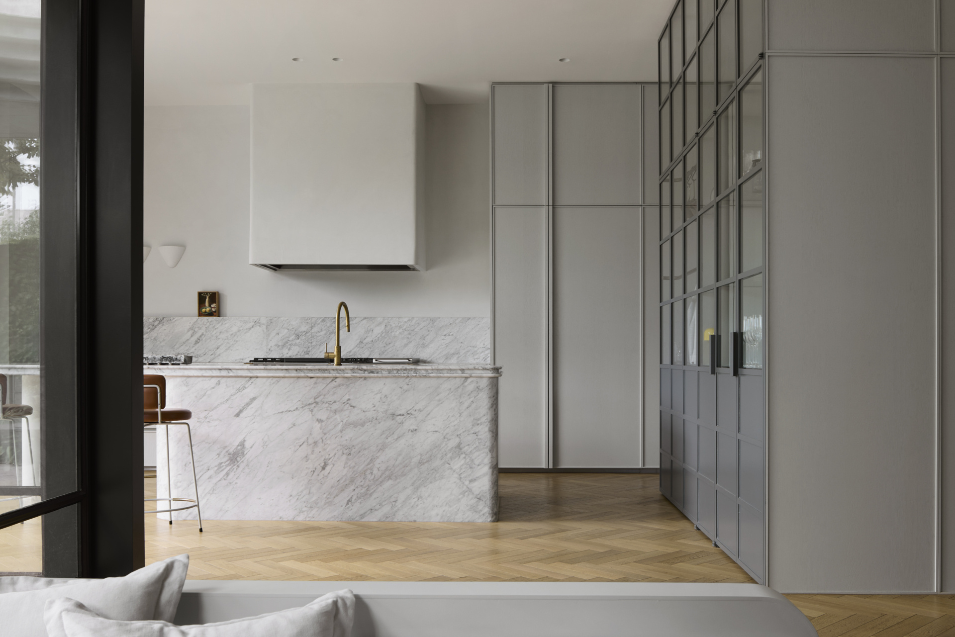 This kitchen finds its rhythm in quiet restraint and considered detail. At its center, a sculptural marble island anchors the space, its creamy veining catching the morning light that filters across pale herringbone floors. Soft grey cabinetry recedes into the background, allowing the island and matching backsplash to take focus. A brass faucet and caramel leather stools bring warmth to the cool palette, while the black-framed glass partition adds structure without closing the space. Framed in black steel and glass, the partition adds structure without closing off the space.