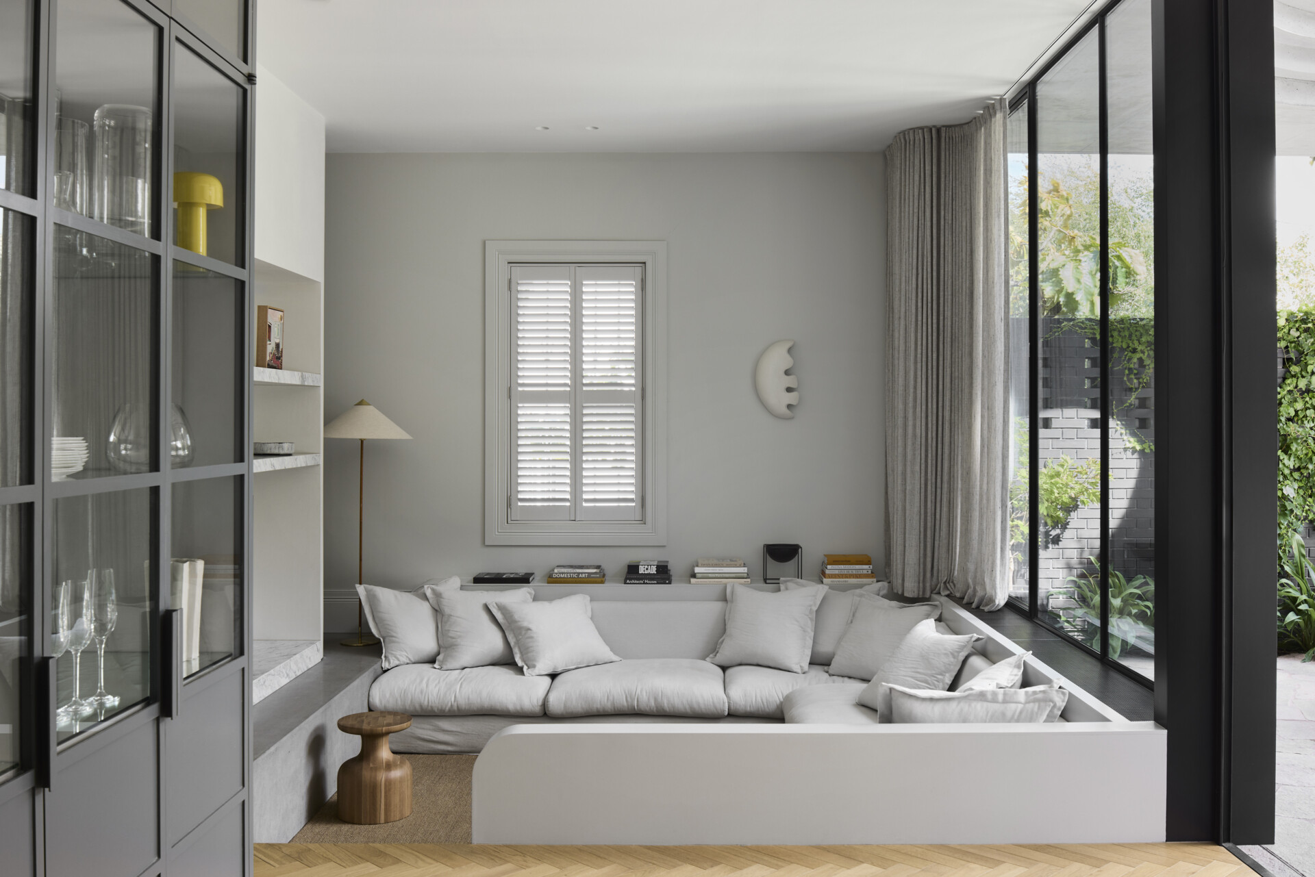 Just beside the kitchen, a sunken lounge offers a quiet shift in pace, its lowered floor creating a sense of enclosure and ease. Soft greys and natural textures define the space, where a deep sectional invites slow afternoons and unhurried conversation. A timber stool adds warmth against the clean lines of the built-in shelving, while full-height glazing draws the garden inward.