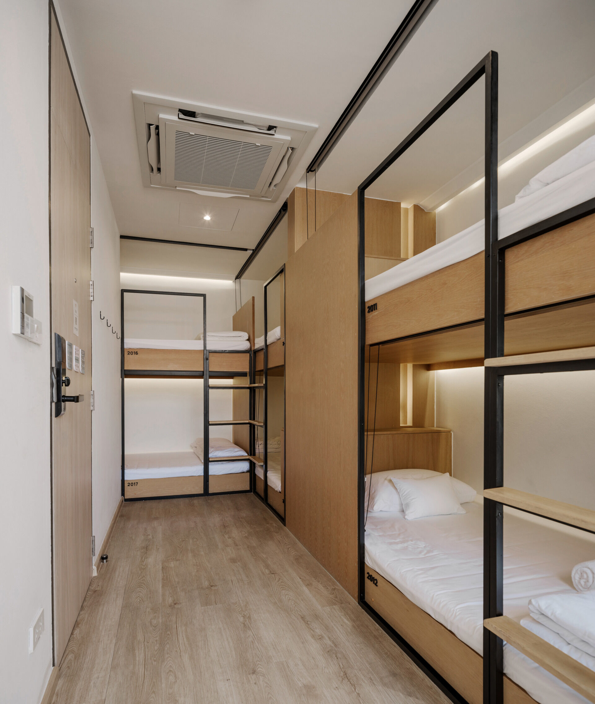 In this modern hostel, guest rooms range from private suites to shared dorm-style spaces with bunk beds, each designed with the same attention to warmth and simplicity found throughout the hostel. Wood accents carry through in the form of open wardrobes, built-in desks, and shelving, maintaining a consistent material language from the common areas. Whether private or shared, each room feels grounded and intentional, offering a calm retreat from the surrounding city.