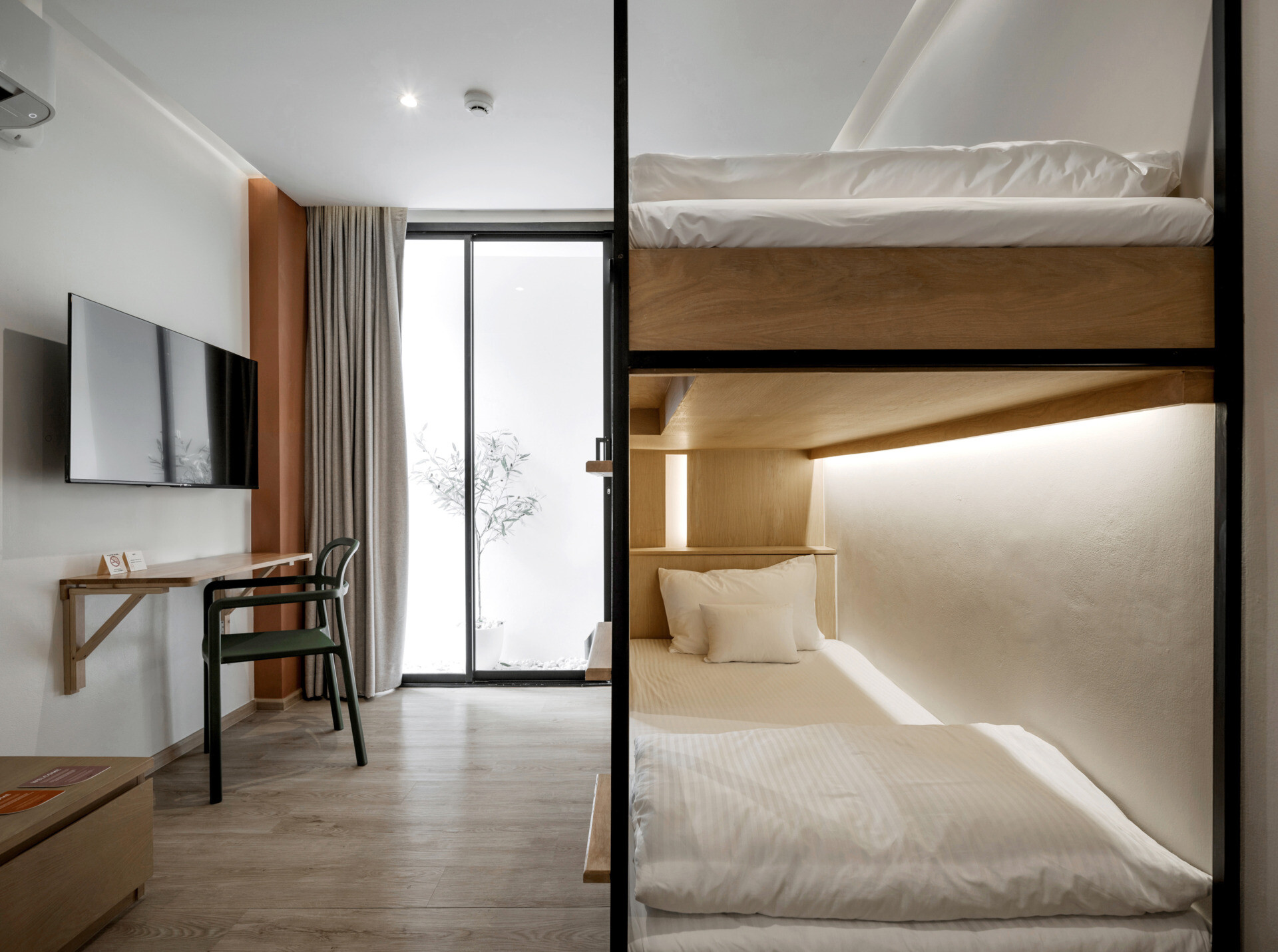 In this modern hostel, guest rooms range from private suites to shared dorm-style spaces with bunk beds, each designed with the same attention to warmth and simplicity found throughout the hostel. Wood accents carry through in the form of open wardrobes, built-in desks, and shelving, maintaining a consistent material language from the common areas. Whether private or shared, each room feels grounded and intentional, offering a calm retreat from the surrounding city.