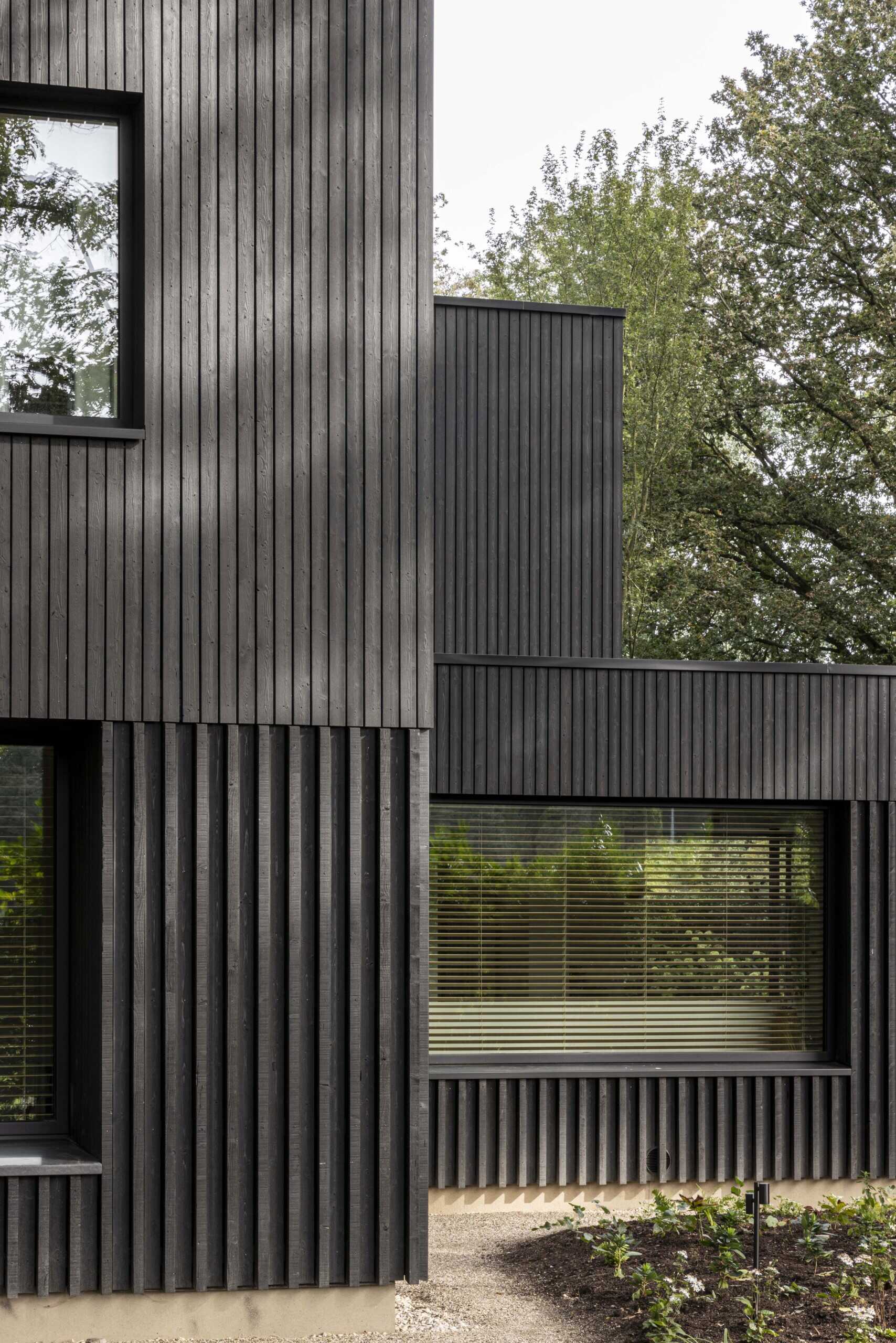 This striking modern home showcases the power of simplicity through its dark wood cladding and clean, rectangular form. Vertical wood slats add texture and rhythm to the facade, while the deep black finish helps the structure blend seamlessly into its natural surroundings. Generously sized windows are thoughtfully positioned to capture views of the landscape and invite daylight into the interior, all while maintaining the home’s strong architectural lines. 