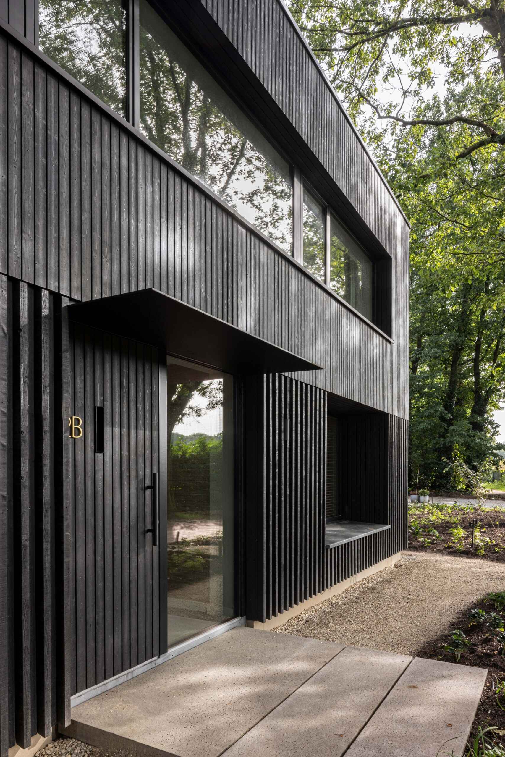 The entryway of this contemporary home is defined by its refined use of dark-stained wood cladding, arranged in narrow vertical strips that give the façade a strong, modern rhythm. The clean lines are softened by the play of natural light and shadow filtering through the surrounding trees, creating a subtle, ever-changing texture on the surface.  A minimalist black canopy extends over the front door, adding a sharp architectural detail while providing a touch of shelter. The path leading to the entrance combines poured concrete slabs and fine gravel, complementing the organic tones of the landscape and guiding visitors toward the recessed doorway with understated elegance.  This close-up view reveals the thoughtful craftsmanship and restrained palette that give the home its calm and confident presence. 