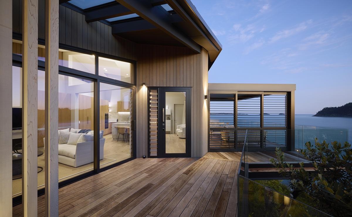 Evening light spills onto this wraparound timber deck, seamlessly connecting indoor living to a sheltered outdoor lounge with sweeping ocean views. Glass balustrades and louvered screens offer openness and privacy, while natural materials keep the home grounded in its stunning coastal setting.