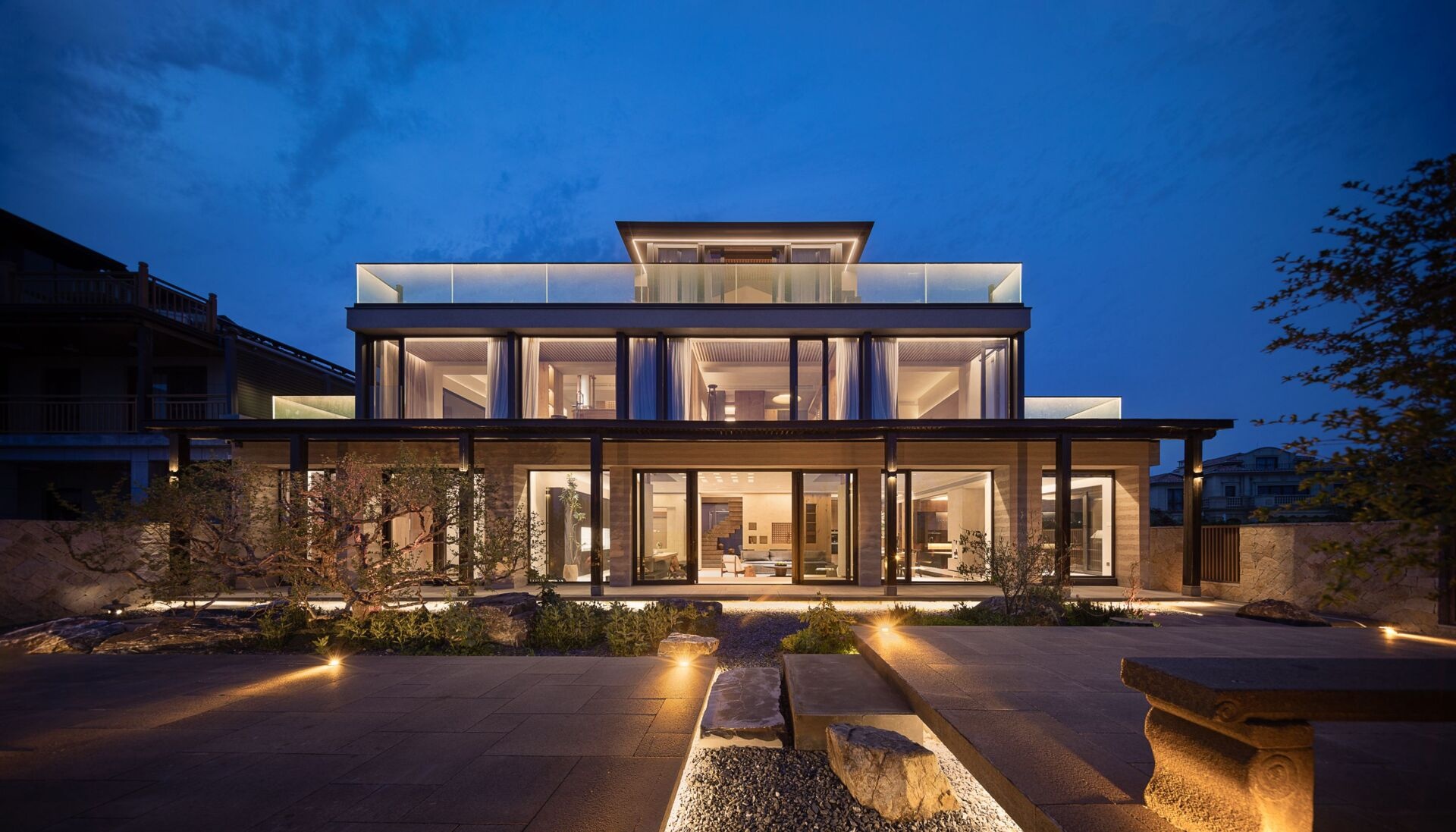 Framed by the soft glow of evening, the rear facade of this home blends sleek lines with warm light to create a welcoming yet elevated atmosphere. Expansive glass panels open the home to its garden, blurring the boundary between indoors and out. The stone pathway, dotted with boulders and low plantings, guides your eye naturally toward the glowing interior. Gentle uplighting and clean architectural symmetry give the whole space a sense of calm balance.