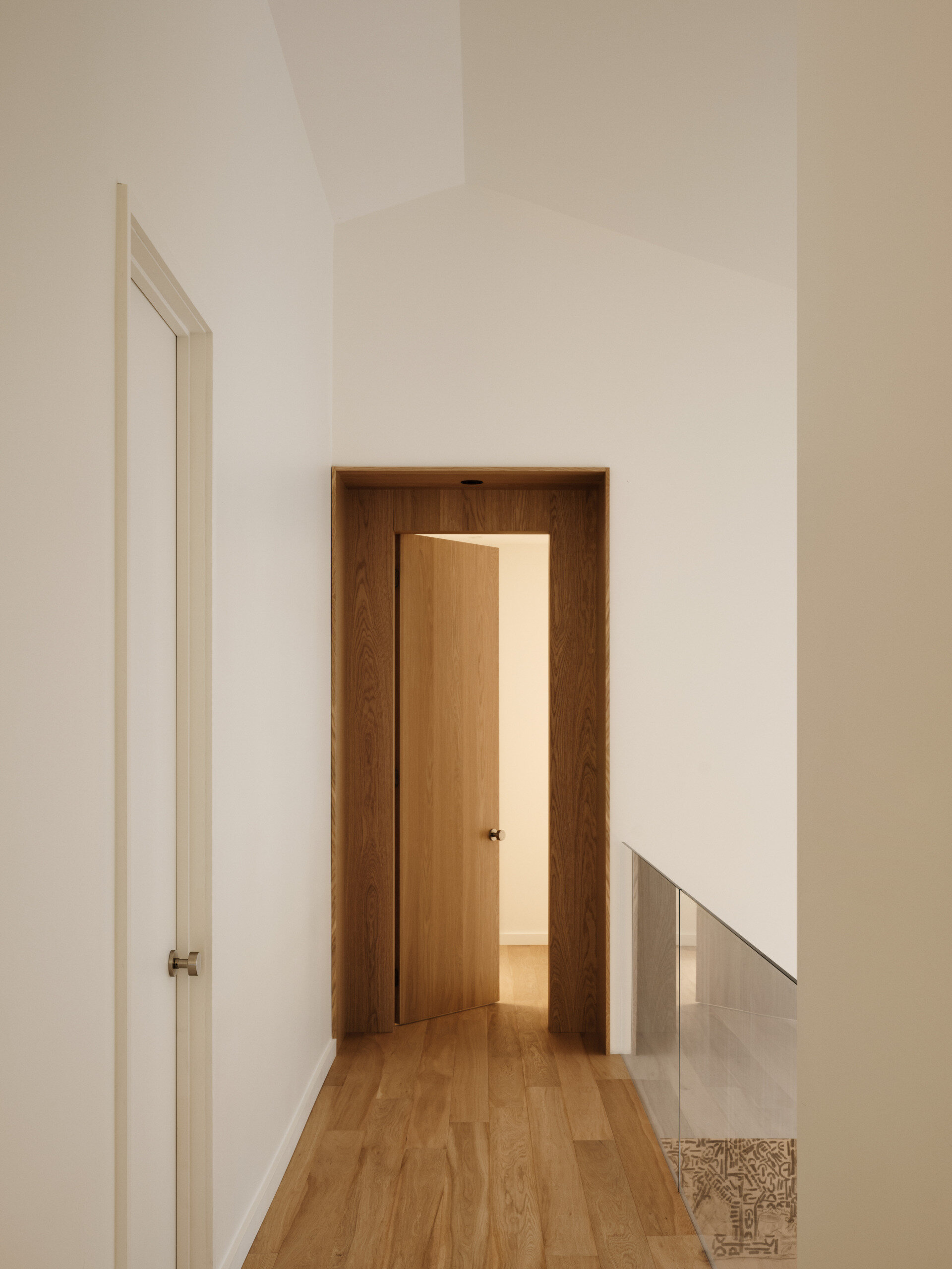 The view of the upstairs hallway leads us through a warm oak-framed threshold, where the door, just slightly ajar, invites curiosity and movement deeper into the home. Framed with intention, the doorway becomes more than just an entry. It feels like a sculptural moment within the architecture. The angled ceiling introduces a subtle architectural rhythm, catching the light and creating depth through shadow, while on the right, an opening and a clear glass balustrade provide a view of the lower level.