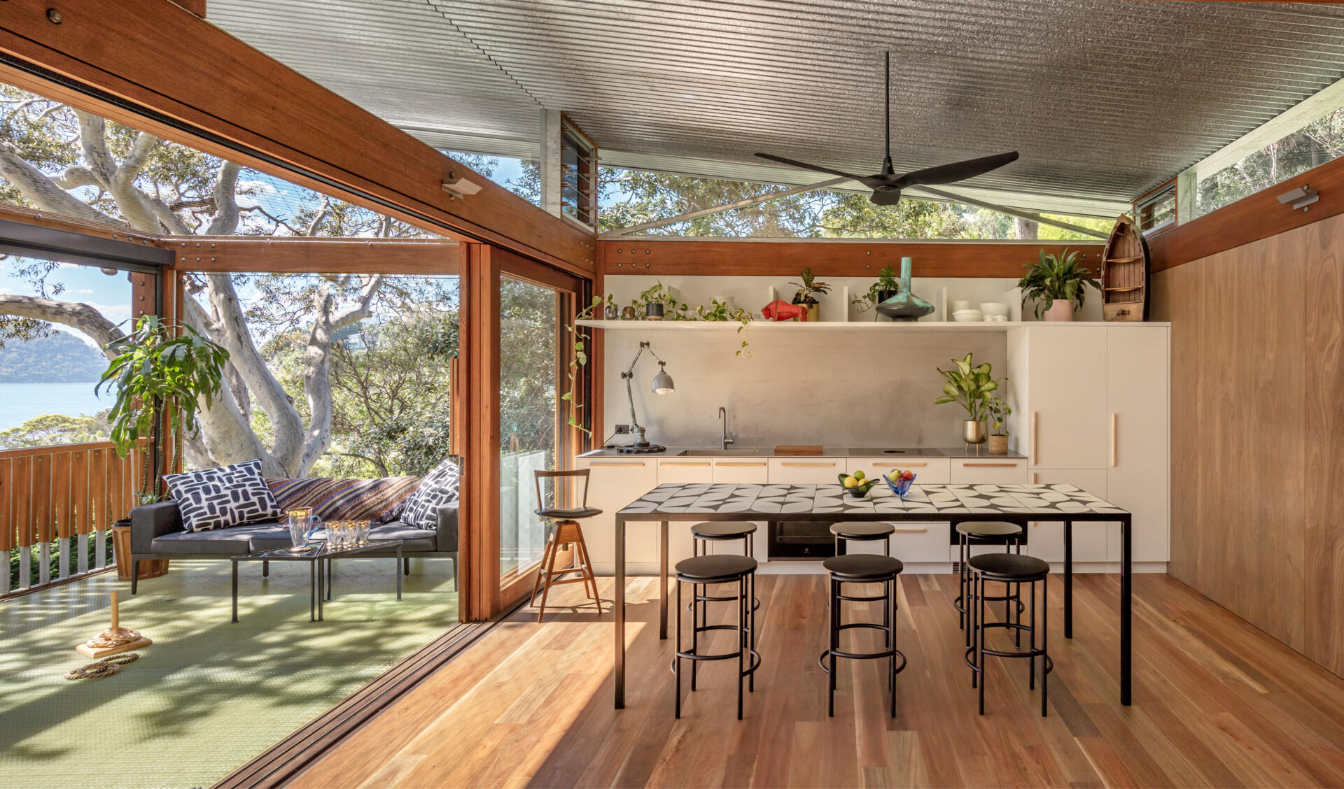 Running along the same outdoor-facing edge, the kitchen and dining area continue to keep things casual. The white cabinetry is modest, with everything within easy reach. Warm wood flooring and frames contrast beautifully with the cool metal ceiling. The tiled island, which also acts as a dining table adds a playful edge, while indoor plants echo the greenery just outside.