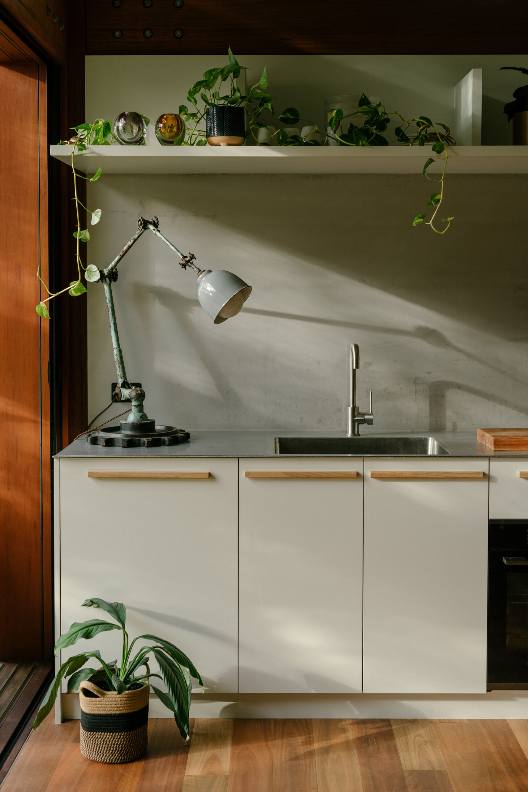 Running along the same outdoor-facing edge, the kitchen and dining area continue to keep things casual. The white cabinetry is modest, with everything within easy reach. Warm wood flooring and frames contrast beautifully with the cool metal ceiling. The tiled island, which also acts as a dining table adds a playful edge, while indoor plants echo the greenery just outside.