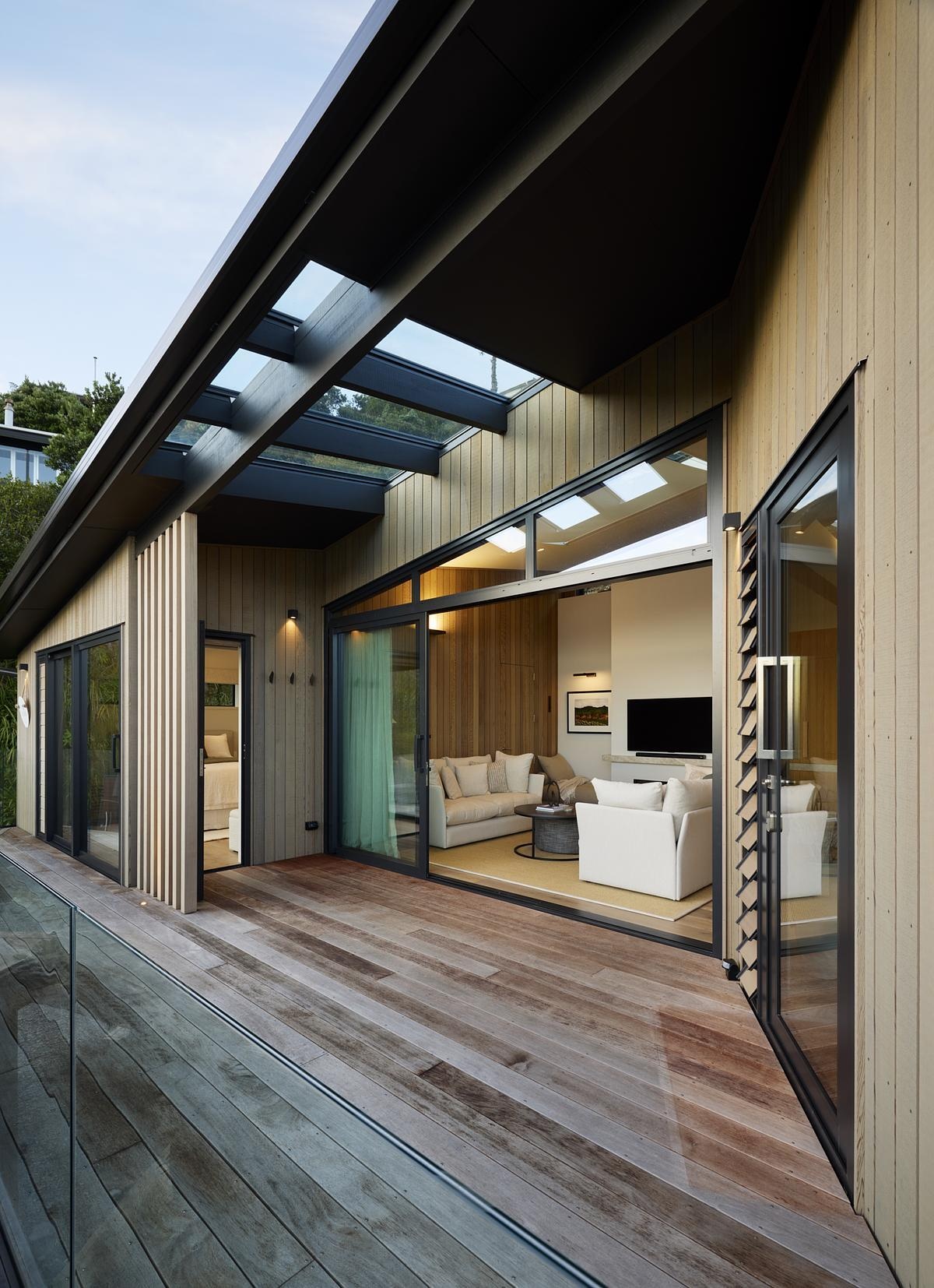 Framed by timber cladding and full-height glass, this cozy living space opens wide to a sun-drenched deck with views beyond. Skylights and sliding doors flood the interior with natural light, while the seamless connection to the outdoors creates an inviting, relaxed atmosphere perfect for coastal living.