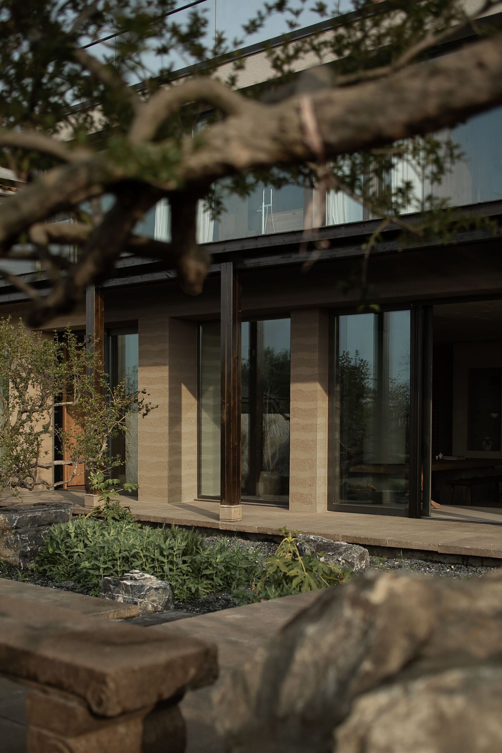 Soft ripples of texture in the rammed earth walls give this home's rear facade a warm, grounded presence that feels deeply tied to the landscape. Floor-to-ceiling glass doors reflect the surrounding garden, letting dappled light spill inside and inviting gentle breezes through the space, while dark wood columns add contrast and rhythm to the architecture.