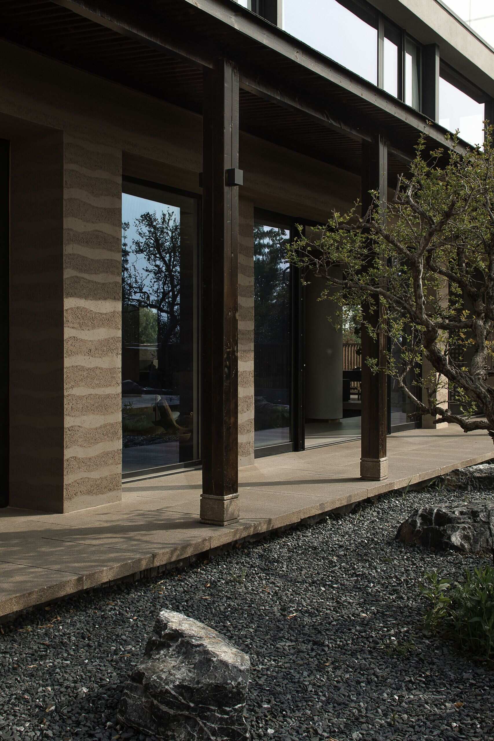 Soft ripples of texture in the rammed earth walls give this home's rear facade a warm, grounded presence that feels deeply tied to the landscape. Floor-to-ceiling glass doors reflect the surrounding garden, letting dappled light spill inside and inviting gentle breezes through the space, while dark wood columns add contrast and rhythm to the architecture.