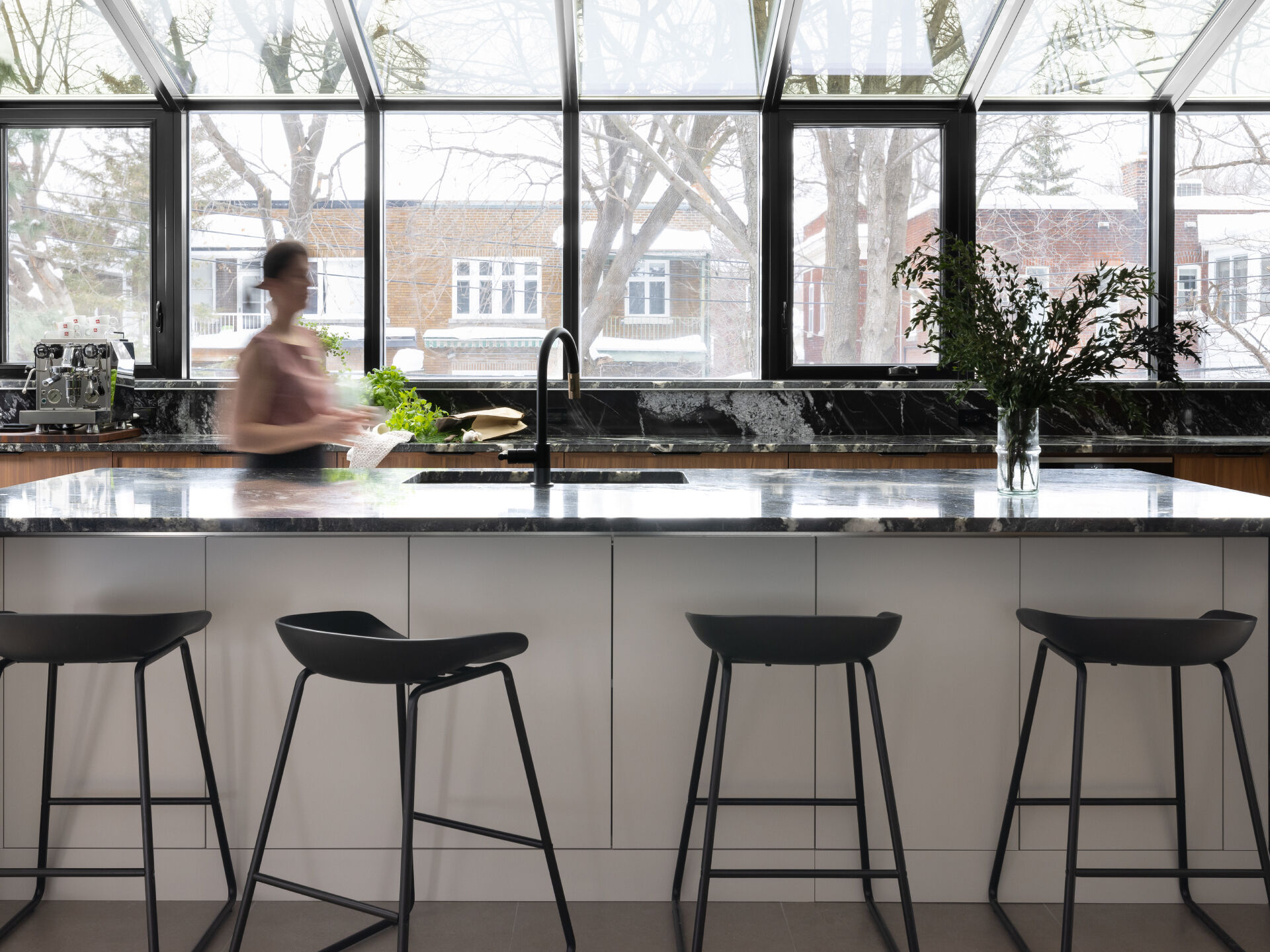 In this modern kitchen, a full glass wall and ceiling frame views of the neighborhood beyond, bathing the space in natural light. Black-framed windows give the room structure without interrupting its openness, while the material palette is rich and deliberate. Bold black marble with expressive white veining wraps the countertops, backsplash, and island waterfall edge, offering visual weight against warm vertical-grain wood cabinetry. A central island anchors the space with clean white drawers and minimalist black stools, while integrated lighting above keeps the mood bright but understated.