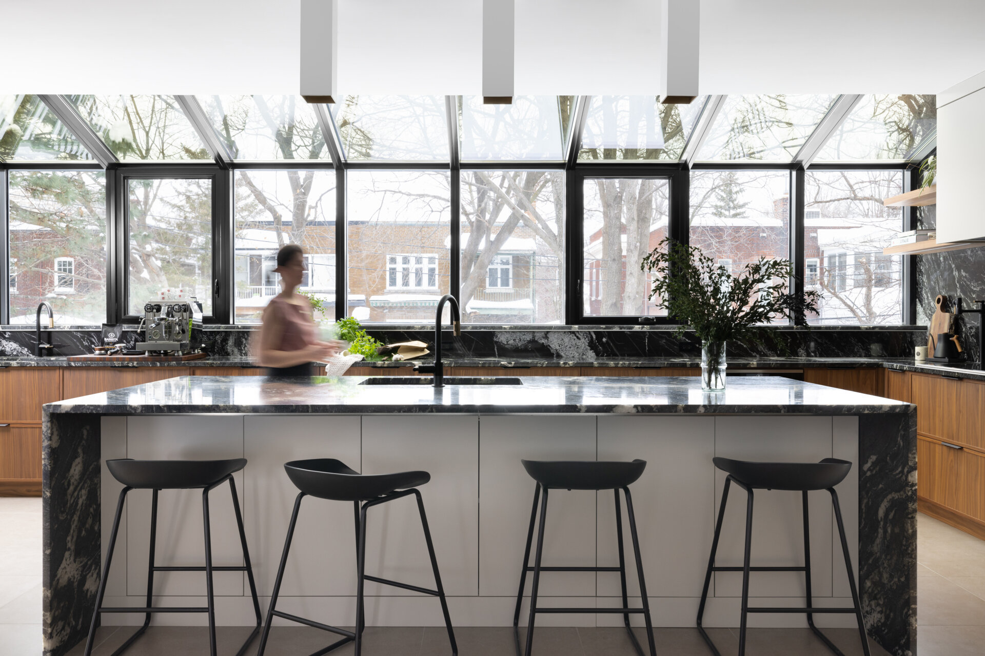 In this modern kitchen, a full glass wall and ceiling frame views of the neighborhood beyond, bathing the space in natural light. Black-framed windows give the room structure without interrupting its openness, while the material palette is rich and deliberate. Bold black marble with expressive white veining wraps the countertops, backsplash, and island waterfall edge, offering visual weight against warm vertical-grain wood cabinetry. A central island anchors the space with clean white drawers and minimalist black stools, while integrated lighting above keeps the mood bright but understated.