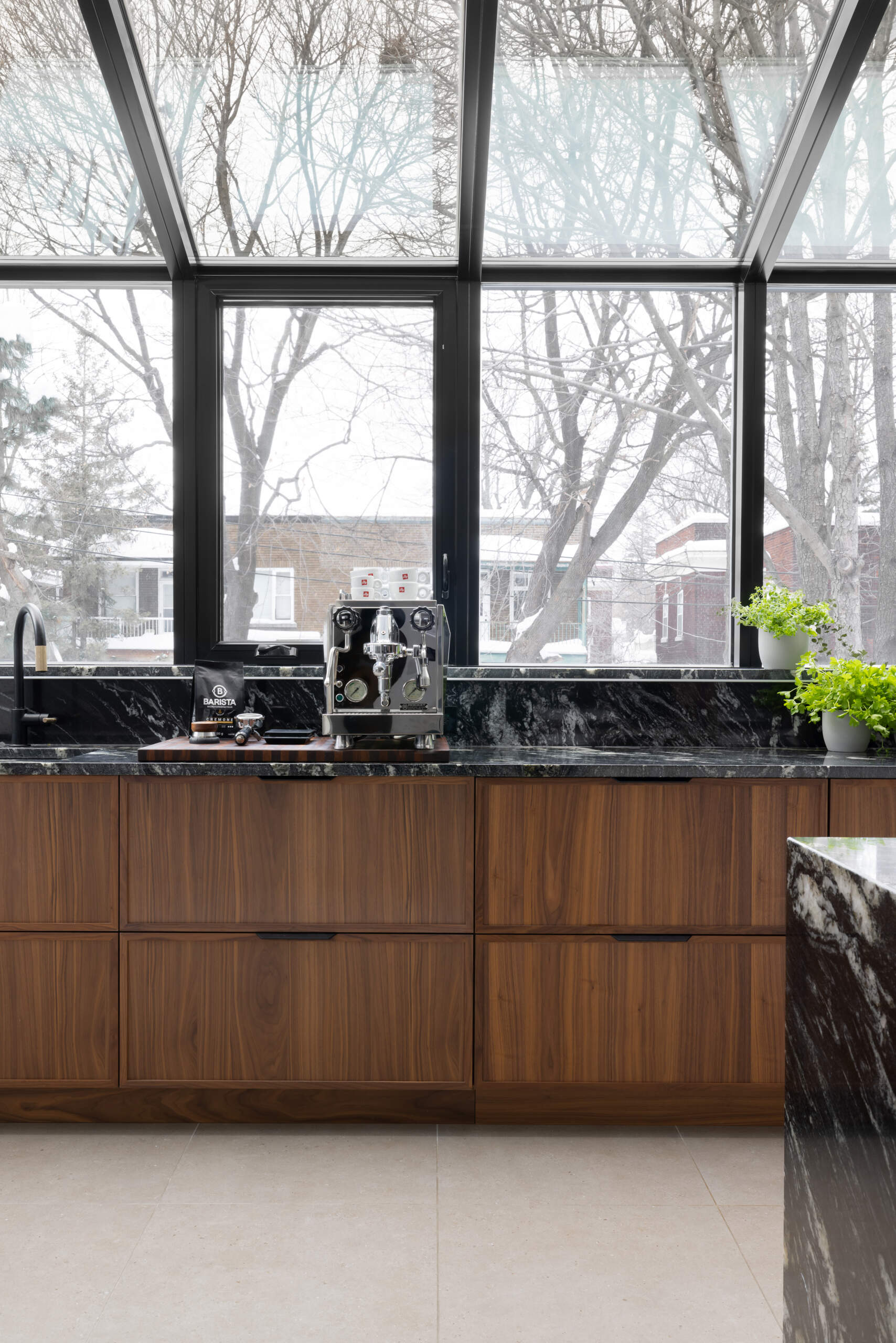 In this modern kitchen, a full glass wall and ceiling frame views of the neighborhood beyond, bathing the space in natural light. Black-framed windows give the room structure without interrupting its openness, while the material palette is rich and deliberate. Bold black marble with expressive white veining wraps the countertops, backsplash, and island waterfall edge, offering visual weight against warm vertical-grain wood cabinetry. A central island anchors the space with clean white drawers and minimalist black stools, while integrated lighting above keeps the mood bright but understated.
