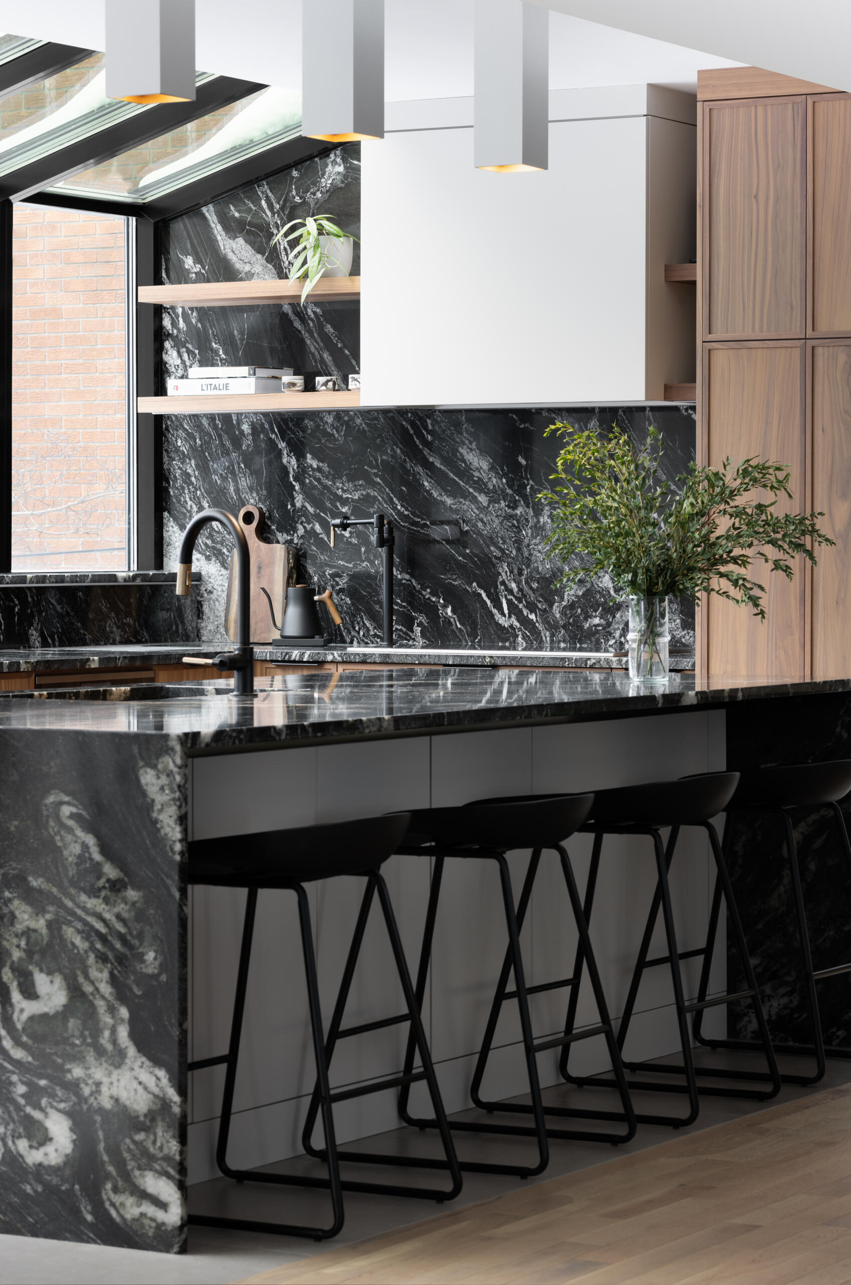 In this modern kitchen, a full glass wall and ceiling frame views of the neighborhood beyond, bathing the space in natural light. Black-framed windows give the room structure without interrupting its openness, while the material palette is rich and deliberate. Bold black marble with expressive white veining wraps the countertops, backsplash, and island waterfall edge, offering visual weight against warm vertical-grain wood cabinetry. A central island anchors the space with clean white drawers and minimalist black stools, while integrated lighting above keeps the mood bright but understated.