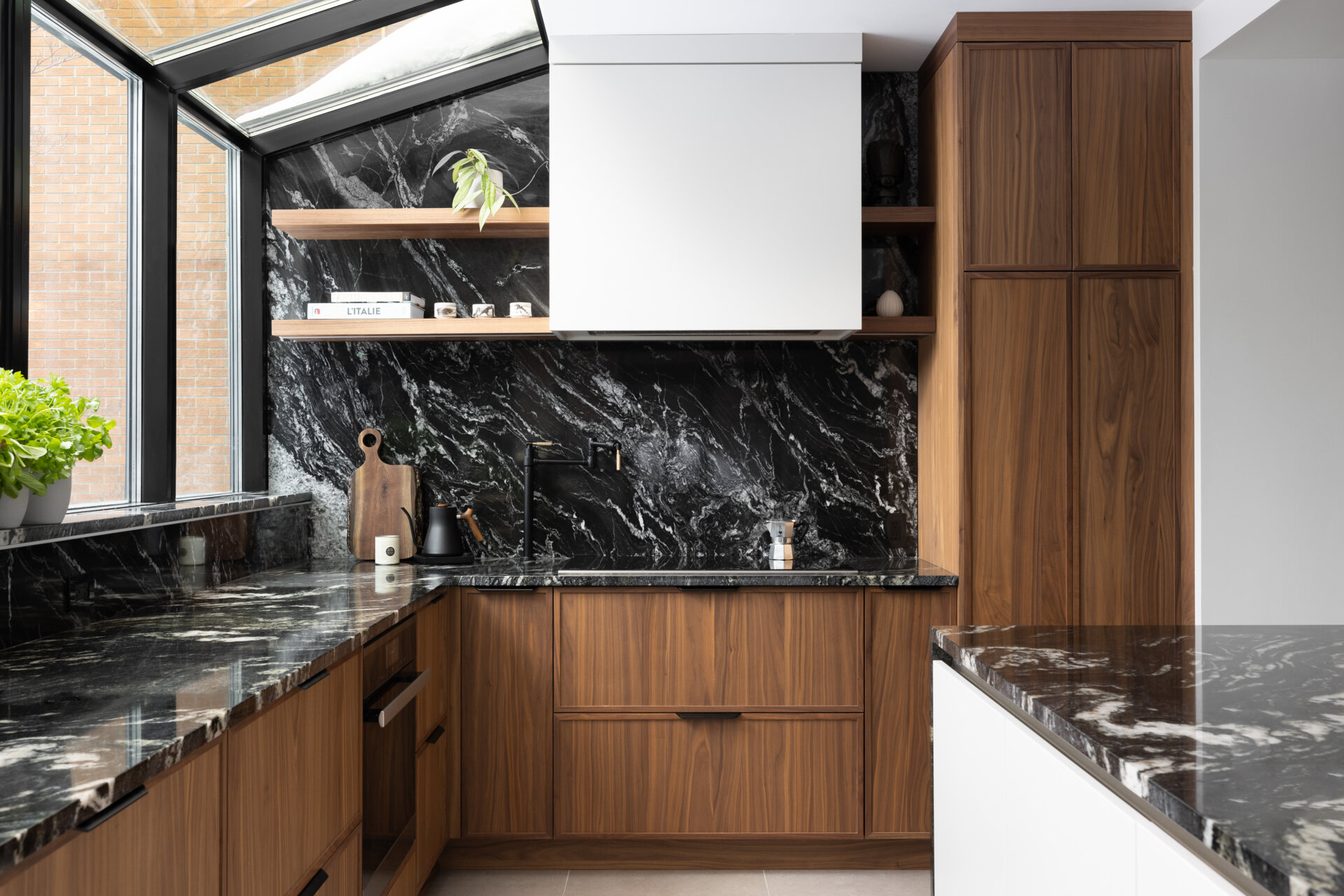 In this modern kitchen, a full glass wall and ceiling frame views of the neighborhood beyond, bathing the space in natural light. Black-framed windows give the room structure without interrupting its openness, while the material palette is rich and deliberate. Bold black marble with expressive white veining wraps the countertops, backsplash, and island waterfall edge, offering visual weight against warm vertical-grain wood cabinetry. A central island anchors the space with clean white drawers and minimalist black stools, while integrated lighting above keeps the mood bright but understated.
