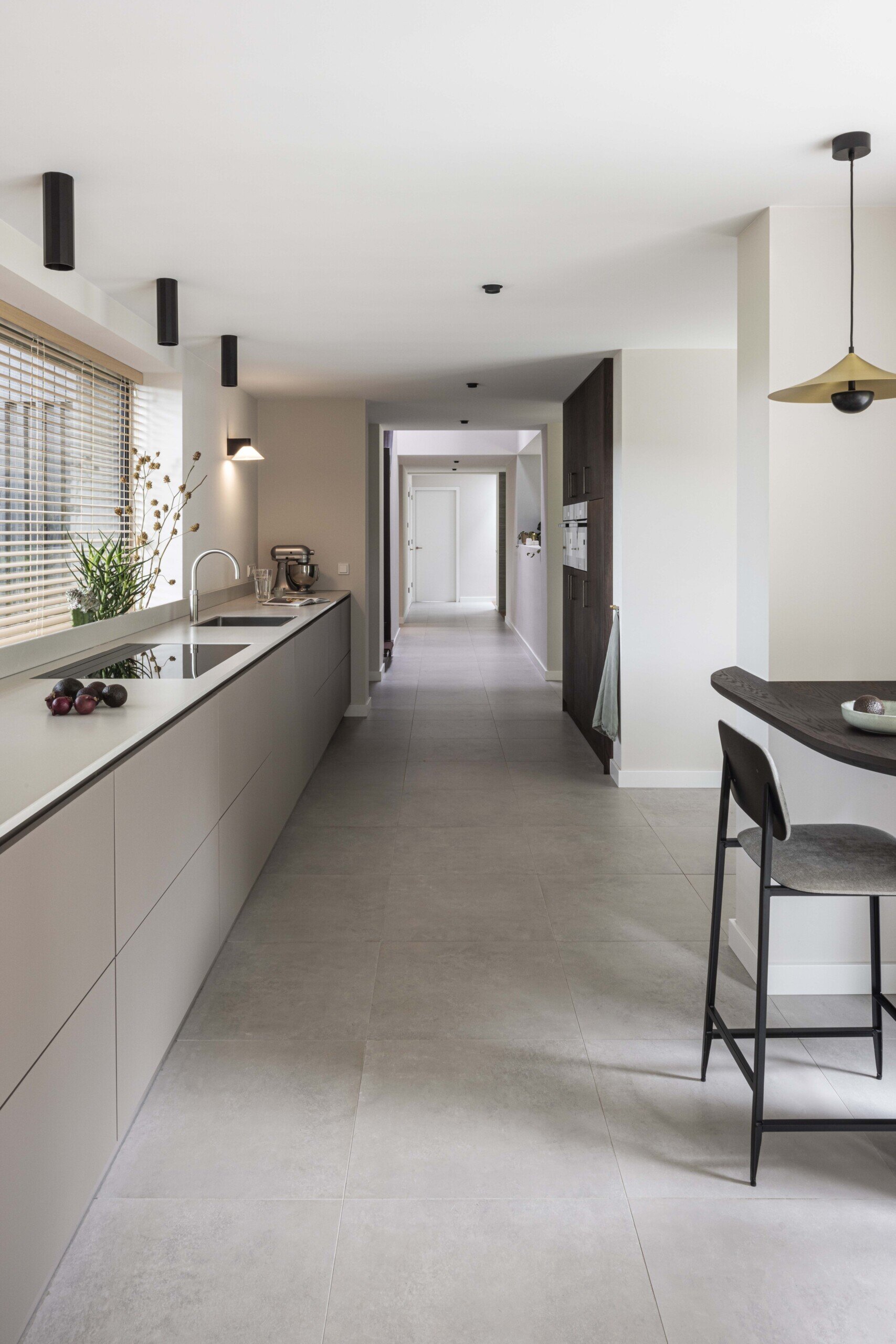 This kitchen combines streamlined design with a calm, inviting atmosphere. Long and linear, the layout emphasizes clarity and function, with soft grey cabinetry running the length of the space and blending seamlessly into the pale tiled floor.  Natural light filters in through a wide window with timber blinds, casting gentle shadows across the quartz benchtop. Touches of greenery and a bowl of dark plums add organic contrast to the otherwise minimal palette. The integrated cooktop and under-mounted sink keep the benchtop visually uninterrupted, enhancing the kitchen’s sleek and uncluttered look.  At the far end, a cozy breakfast bar introduces a note of warmth with its dark wood counter and simple bar stool. Overhead, a sculptural pendant light offers just enough character without disturbing the overall sense of balance and restraint.