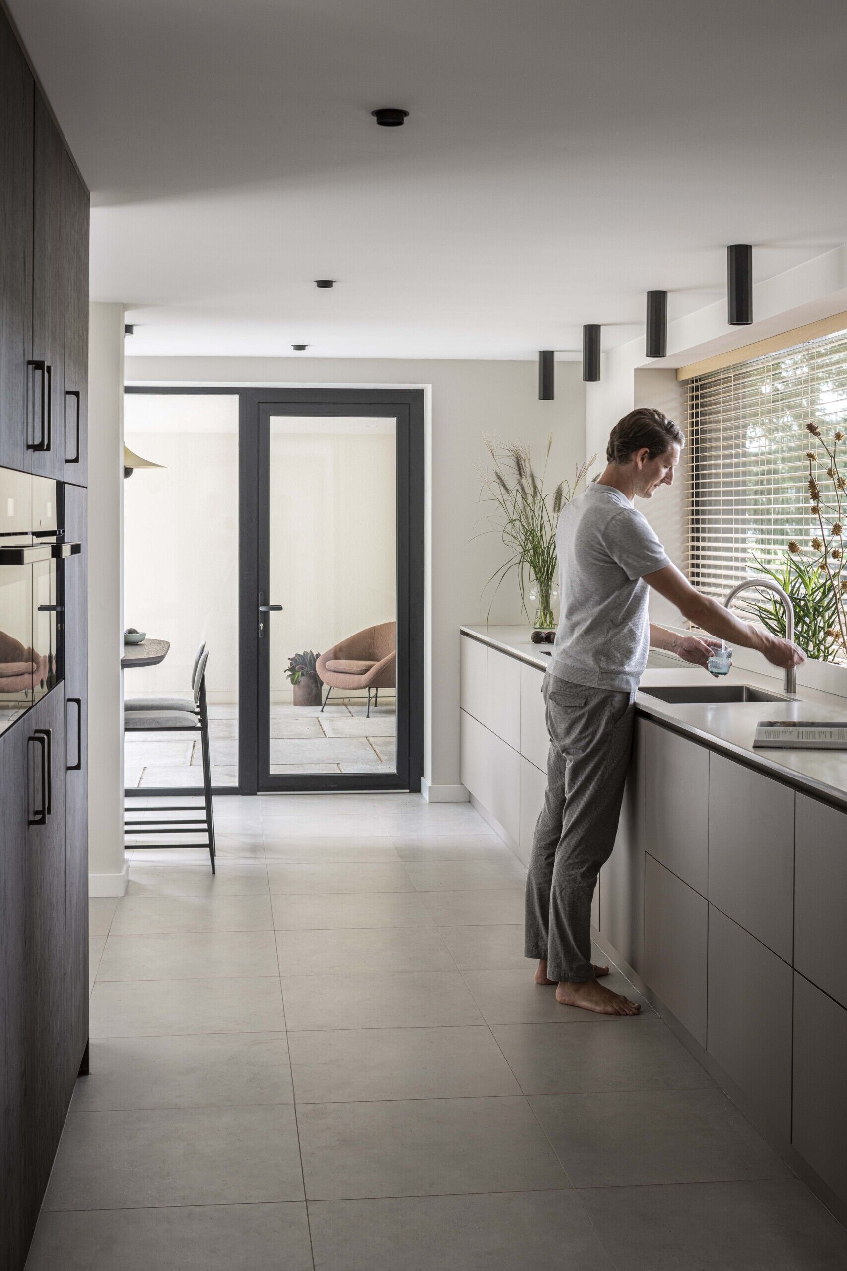 This kitchen combines streamlined design with a calm, inviting atmosphere. Long and linear, the layout emphasizes clarity and function, with soft grey cabinetry running the length of the space and blending seamlessly into the pale tiled floor.  Natural light filters in through a wide window with timber blinds, casting gentle shadows across the quartz benchtop. Touches of greenery and a bowl of dark plums add organic contrast to the otherwise minimal palette. The integrated cooktop and under-mounted sink keep the benchtop visually uninterrupted, enhancing the kitchen’s sleek and uncluttered look.  At the far end, a cozy breakfast bar introduces a note of warmth with its dark wood counter and simple bar stool. Overhead, a sculptural pendant light offers just enough character without disturbing the overall sense of balance and restraint.