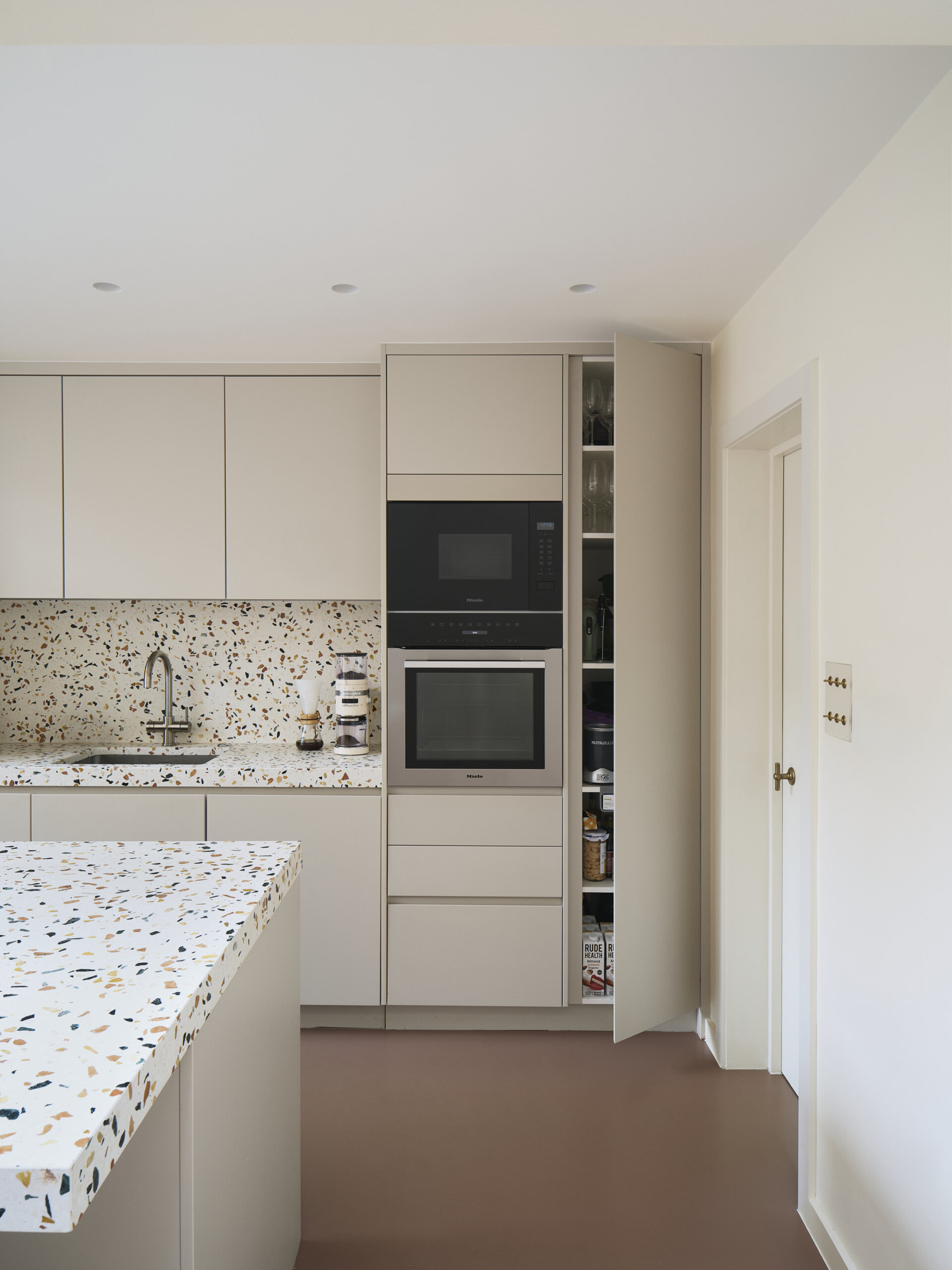 The kitchen features a clean, integrated design with hardware-free cabinetry that complements the home’s minimalist material palette. Terrazzo countertops introduce color and variation, adding a subtle sense of play. A central island anchors the room, providing ample space for cooking and casual dining while preserving open views toward the dining area and terrace.