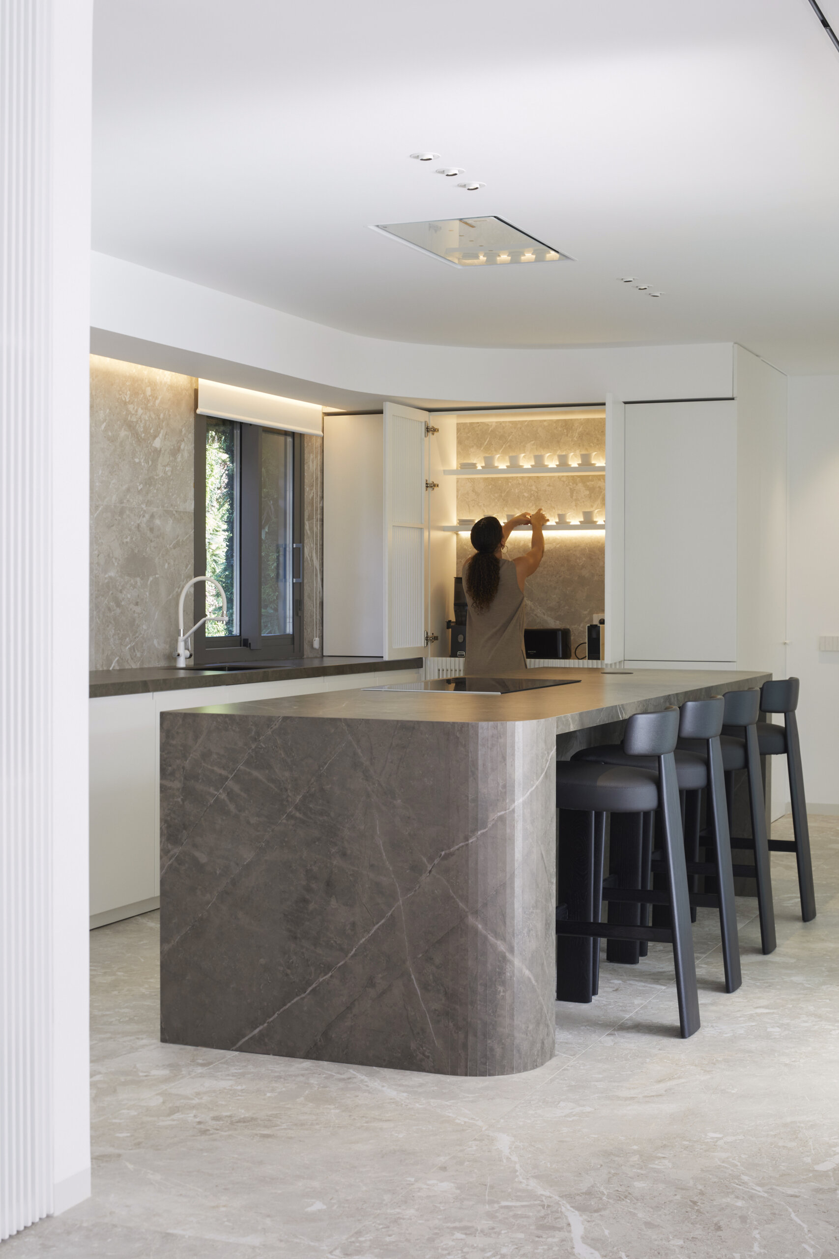 The kitchen balances sculptural form with everyday function. The island, clad in a rich gray stone with bold veining and a softly curved edge, grounds the space while offering plenty of room for casual meals or conversation over coffee. Matte black stools line up neatly, their solid presence echoing the clean lines found throughout. Light filters in through a window, bouncing gently off the stone surfaces and white cabinetry.