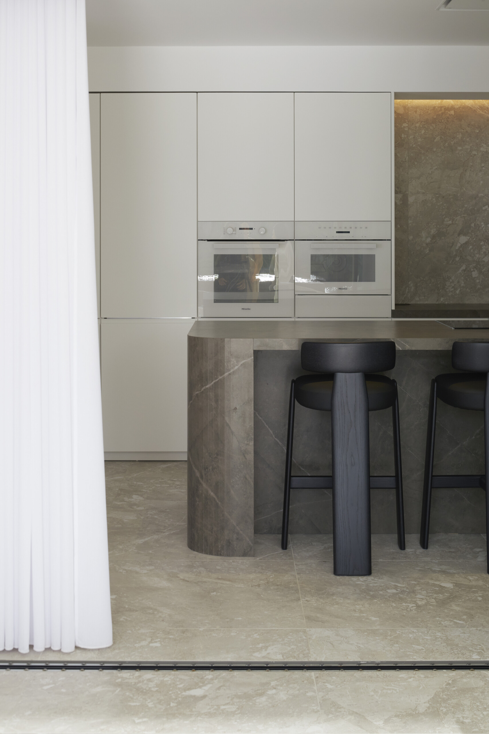 The kitchen balances sculptural form with everyday function. A curved-edge stone island anchors the space, with matte black stools and a hidden pantry moment adding depth to its clean, purposeful rhythm.