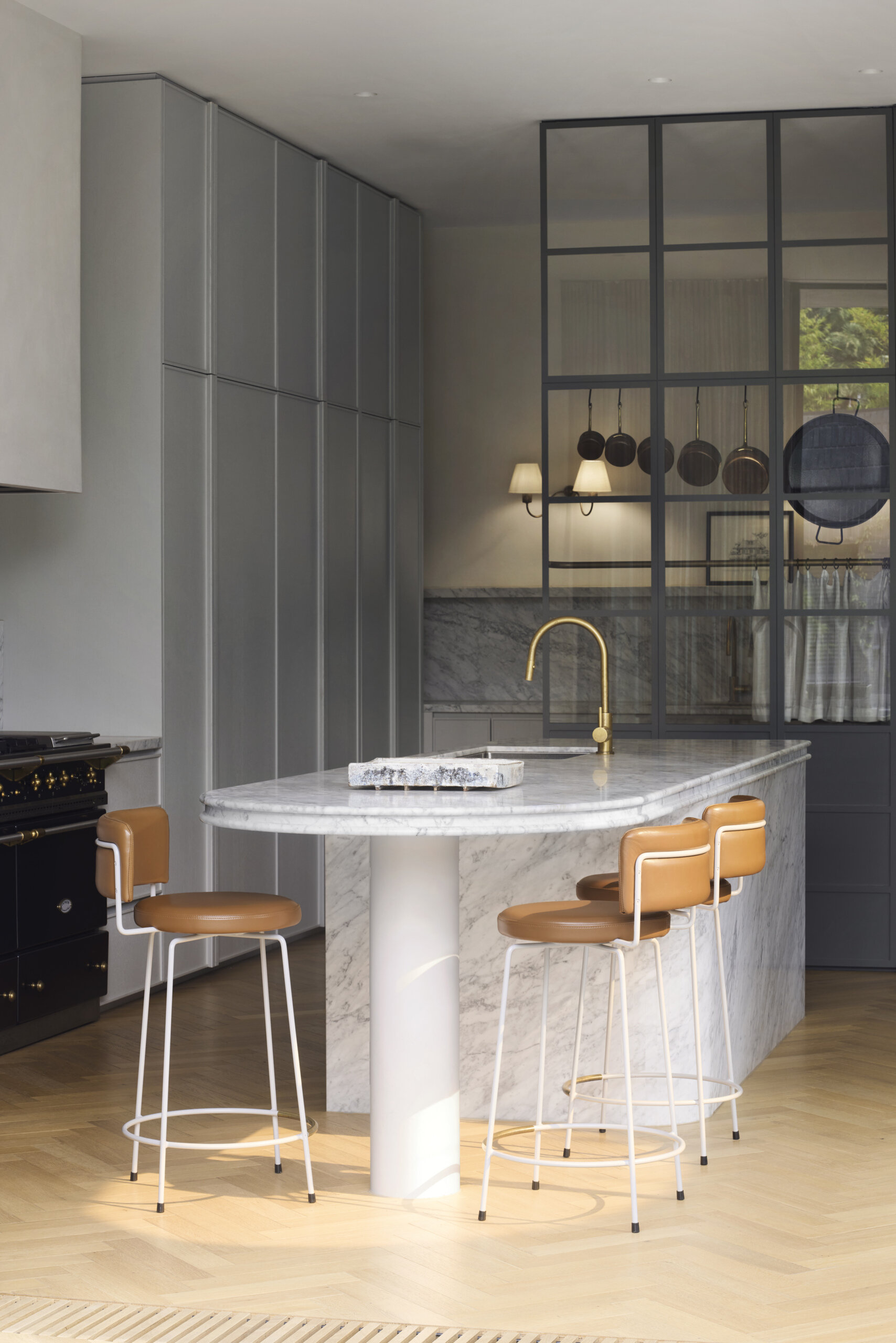 This kitchen finds its rhythm in quiet restraint and considered detail. At its center, a sculptural marble island anchors the space, its creamy veining catching the morning light that filters across pale herringbone floors. Soft grey cabinetry recedes into the background, allowing the island and matching backsplash to take focus. A brass faucet and caramel leather stools bring warmth to the cool palette, while the black-framed glass partition adds structure without closing the space. Framed in black steel and glass, the partition adds structure without closing off the space.