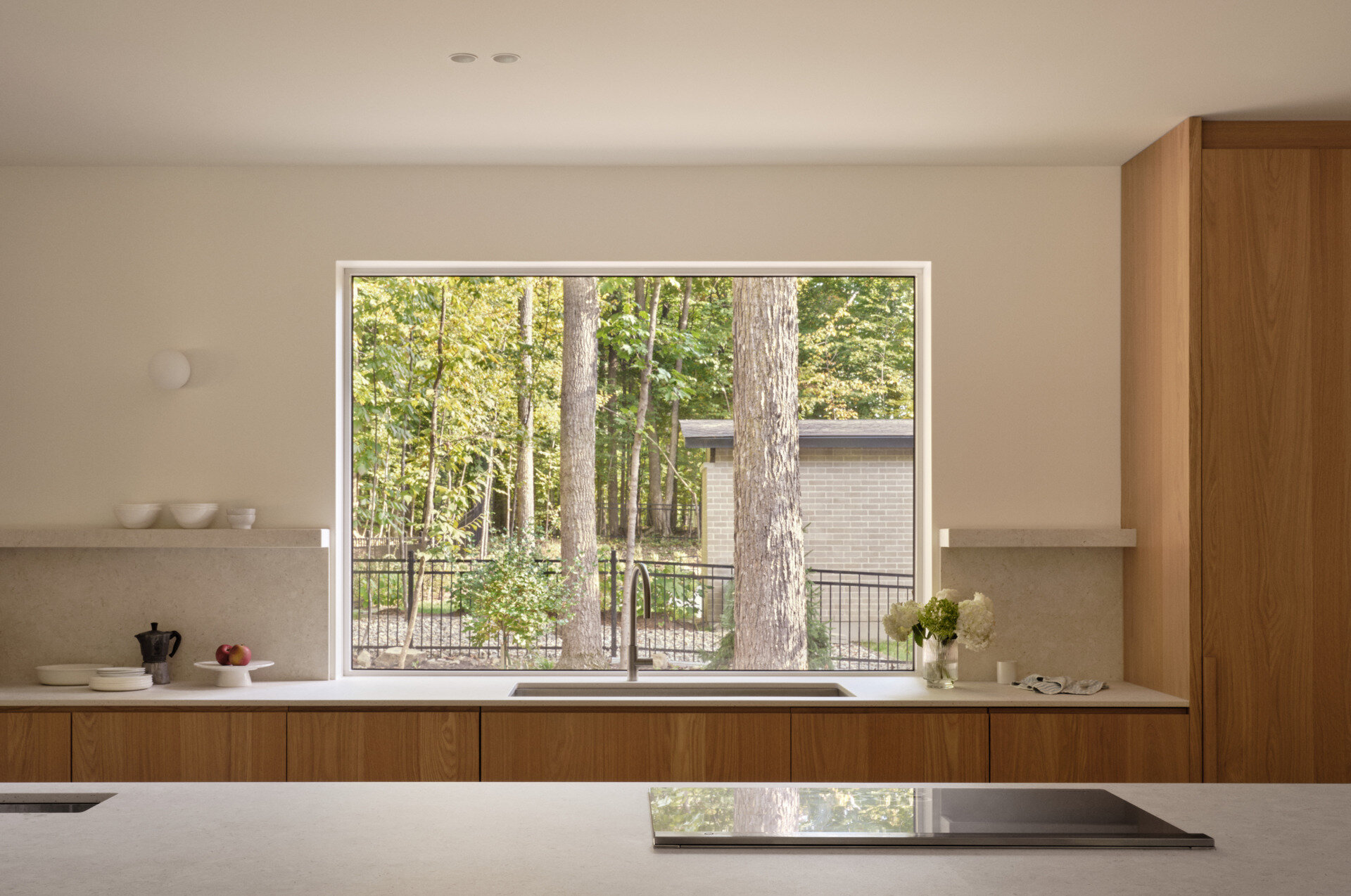The kitchen blends quiet elegance with smart functionality. A wide picture window frames the forest, turning light and landscape into a living artwork. Natural wood cabinetry, pale stone surfaces, and hidden appliances keep the space clean and calm. A built-in coffee nook adds warmth, while the nearby dining area offers a cozy, wood-lined corner for slow mornings.