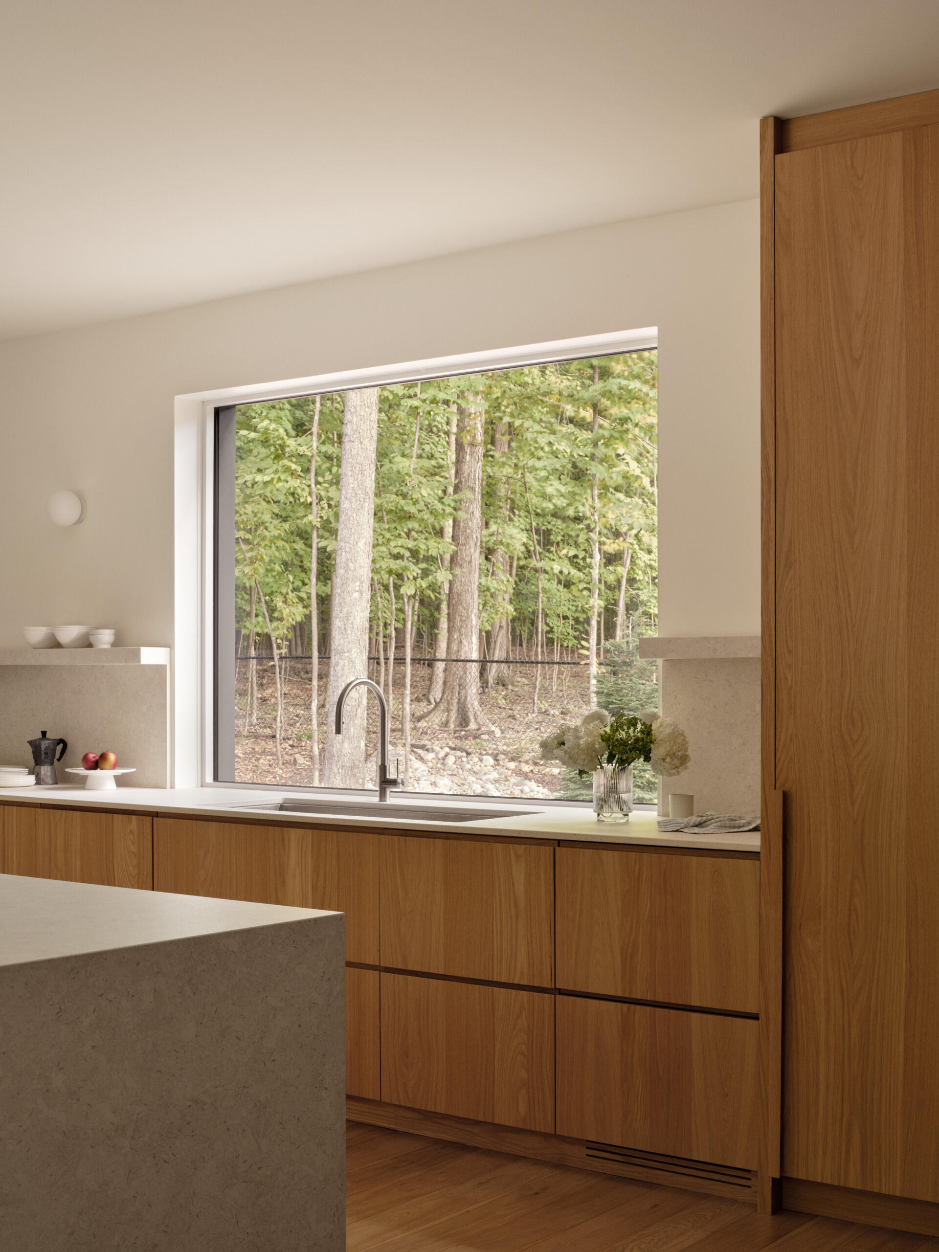 The kitchen blends quiet elegance with smart functionality. A wide picture window frames the forest, turning light and landscape into a living artwork. Natural wood cabinetry, pale stone surfaces, and hidden appliances keep the space clean and calm. A built-in coffee nook adds warmth, while the nearby dining area offers a cozy, wood-lined corner for slow mornings.