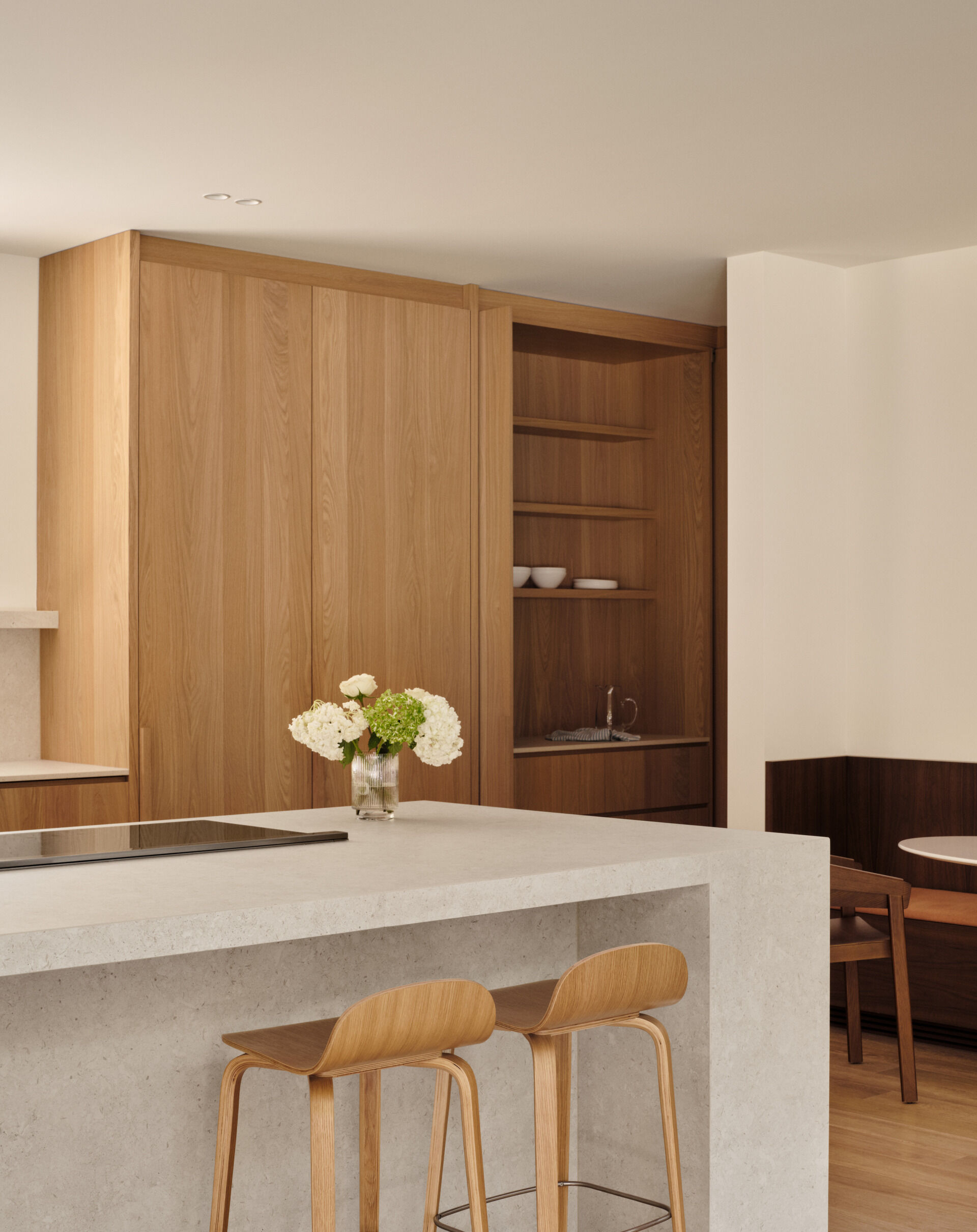 The kitchen blends quiet elegance with smart functionality. A wide picture window frames the forest, turning light and landscape into a living artwork. Natural wood cabinetry, pale stone surfaces, and hidden appliances keep the space clean and calm. A built-in coffee nook adds warmth, while the nearby dining area offers a cozy, wood-lined corner for slow mornings.