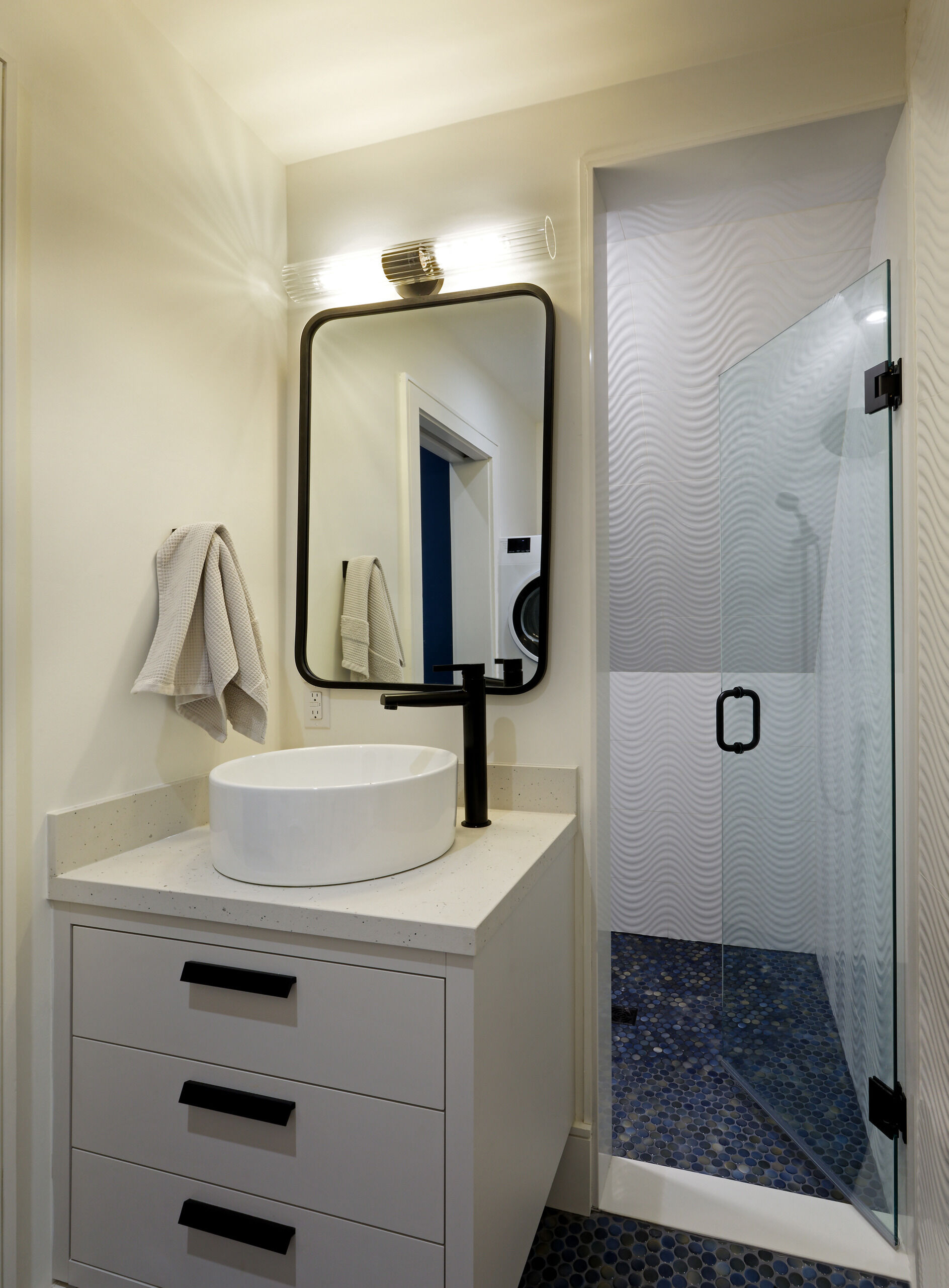 This bathroom is compact but full of texture. A round vessel sink rests on a drawer-fronted vanity, paired with matte black fixtures and a rounded rectangle mirror. The shower enclosure is lined with wavy tiles, and both the shower floor and bathroom floor sparkle with small, gem-like penny tiles that catch the light and add a touch of quiet glamour.